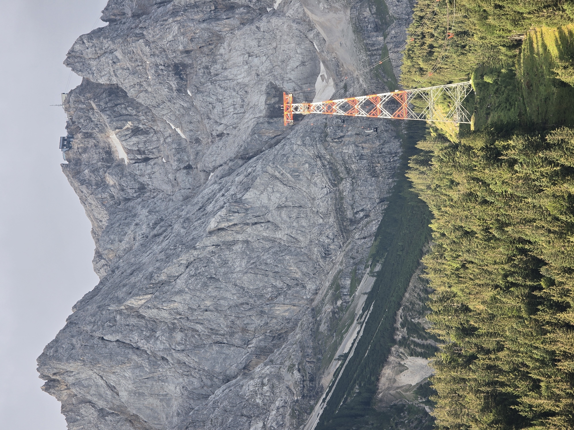 The image captures the Zugspitze cable car pylon, a prominent landmark in Grainau, Germany. The pylon, a striking red and white lattice structure, stands tall amidst a dense, evergreen forest at the base of a colossal, grey, rocky mountain range. The mountain's rugged facade is deeply fissured, with patches of snow clinging to its upper reaches. At the summit of the mountain, a building complex and a communications tower are visible. Several cable cars, small dark pods suspended from wires, are seen in motion, ascending towards the peak. The scene is bathed in natural light, suggesting it was taken during the day, with the sun casting shadows and highlighting the textures of the landscape. The environment is pristine and mountainous, typical of the Bavarian Alps. No people are visible in the image.