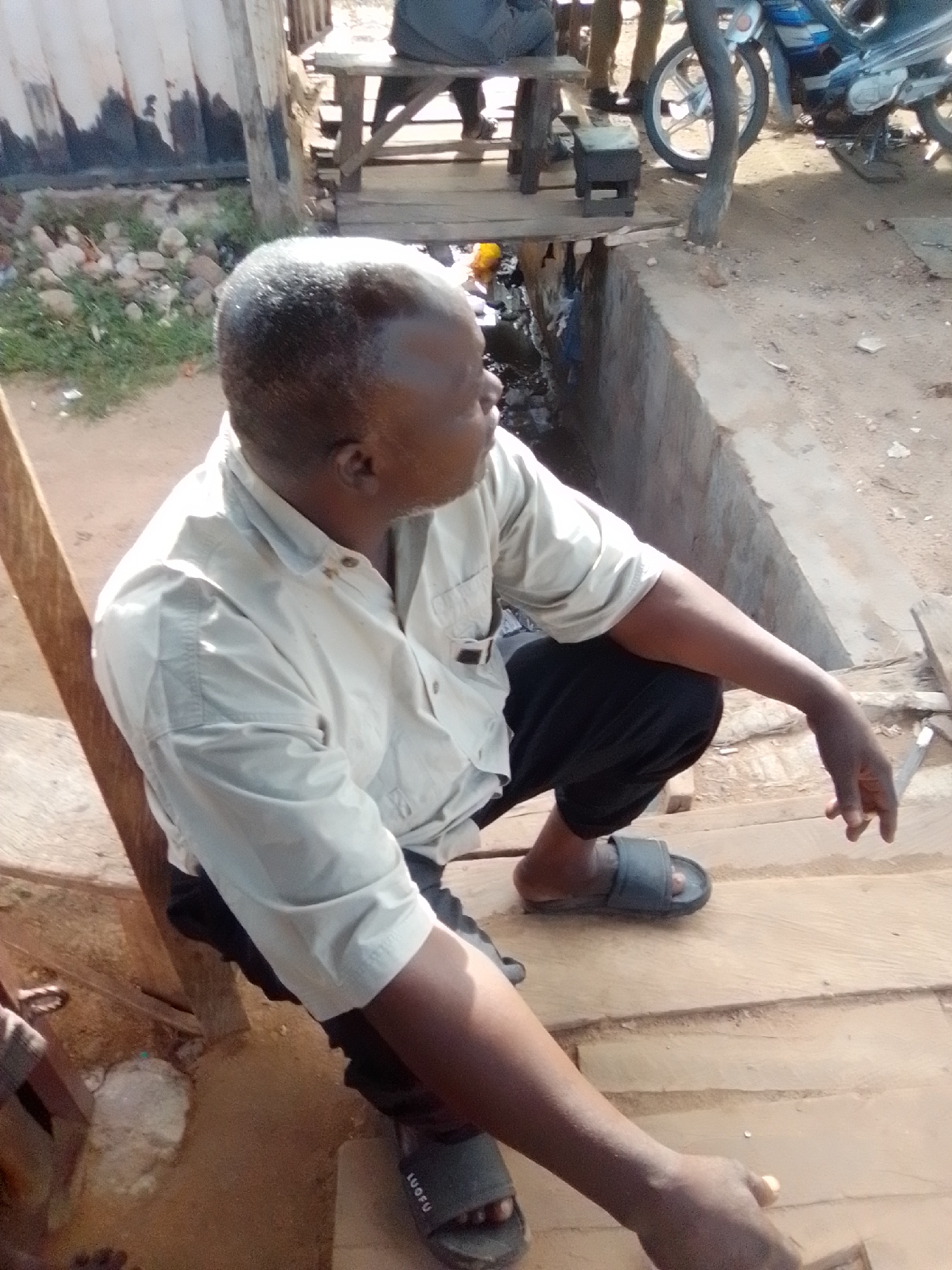 A medium shot from a slightly high angle captures a dark-skinned man with a shaved head, wearing a light-colored button-down shirt and dark pants, seated on wooden steps in what appears to be Pantame, Nigeria. He is looking to his right, with his right arm resting on his bent right leg, and his left hand dangling near the wooden surface. The man is wearing dark-colored slides on his feet, with one foot visible on a lower step.
The environment is rustic and outdoors. To the left of the man, there's a corrugated metal structure with some white painted sections. Adjacent to this structure, there's green grass and scattered rocks. Behind the man, the wooden steps continue upwards, leading to a wooden table. Partially visible behind the table, a blue and white motorcycle is parked. The ground is dirt, with some scattered debris. The lighting suggests it is daytime, with shadows indicating the sun is present. The overall impression is one of an outdoor working area or a casual outdoor gathering space.