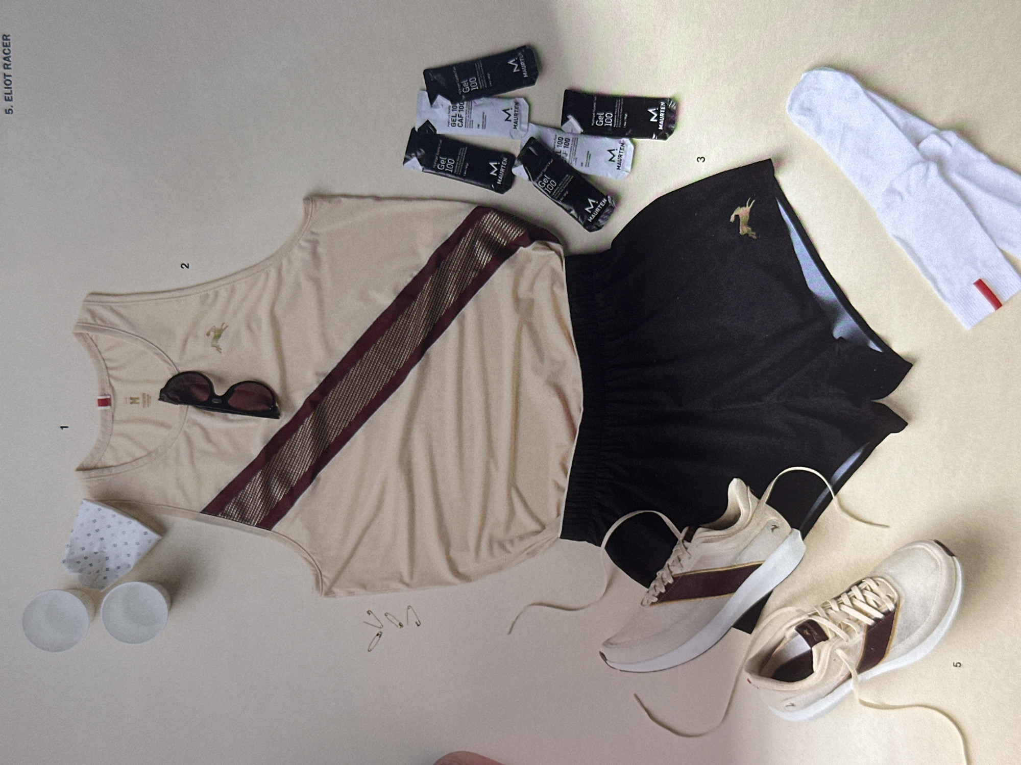The image presents a flat lay of athletic apparel and accessories on a light beige surface, labeled with numerical markers from 1 to 5. At the top left, denoted by the number 1, is a beige tank top with a dark burgundy stripe running diagonally across the front. A pair of sunglasses rests on top of the tank, next to a small patterned cloth and two white cups. Positioned near the upper-center, marked with the number 2, is a cluster of "MAURTEN" energy gel packets. Adjacent to these, indicated by the number 3, are black running shorts featuring a golden antelope emblem, complemented by a pair of white athletic socks with a red stripe at the top. At the bottom right, labeled as number 5, are two pairs of beige running shoes with burgundy stripes. Three safety pins are scattered between the shirt and shoes. The lighting is soft and diffused, creating a clean and minimalist aesthetic. In the top left corner the text "5. ELIOT RACER" is visible.