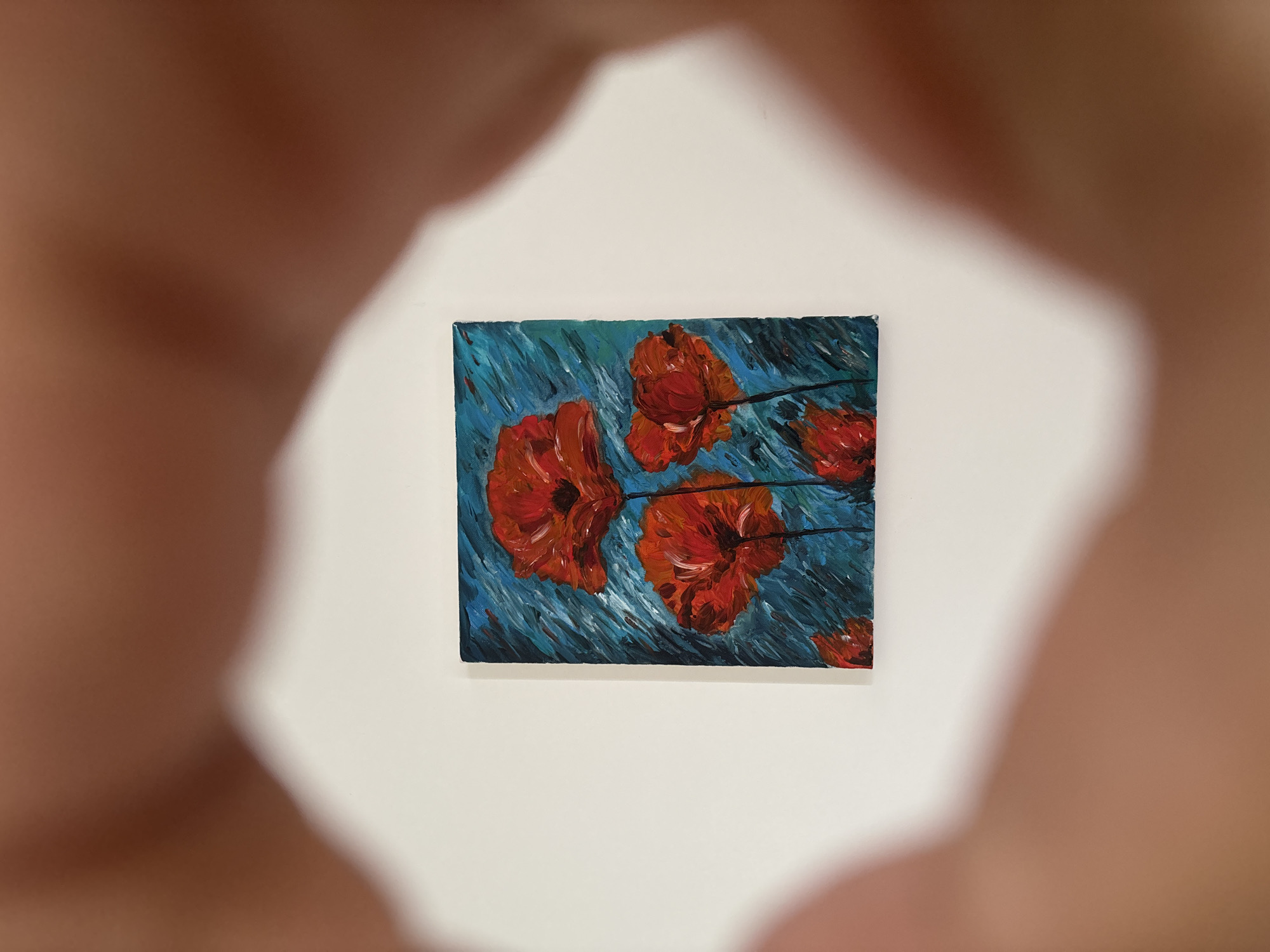 A small, rectangular painting of four red poppies is centered on a white background. The poppies are depicted with thick, textured brushstrokes in shades of red and orange, with dark centers. The background is a vibrant turquoise and blue, with vertical and diagonal strokes that suggest movement. The painting is framed by the blurred-out hands of a person, which create a circular opening through which the artwork is viewed. The hands are a light brown color and appear to be resting on the white surface. The lighting is soft and diffused, casting a gentle glow on the scene. There are no discernible features of any city or specific location.