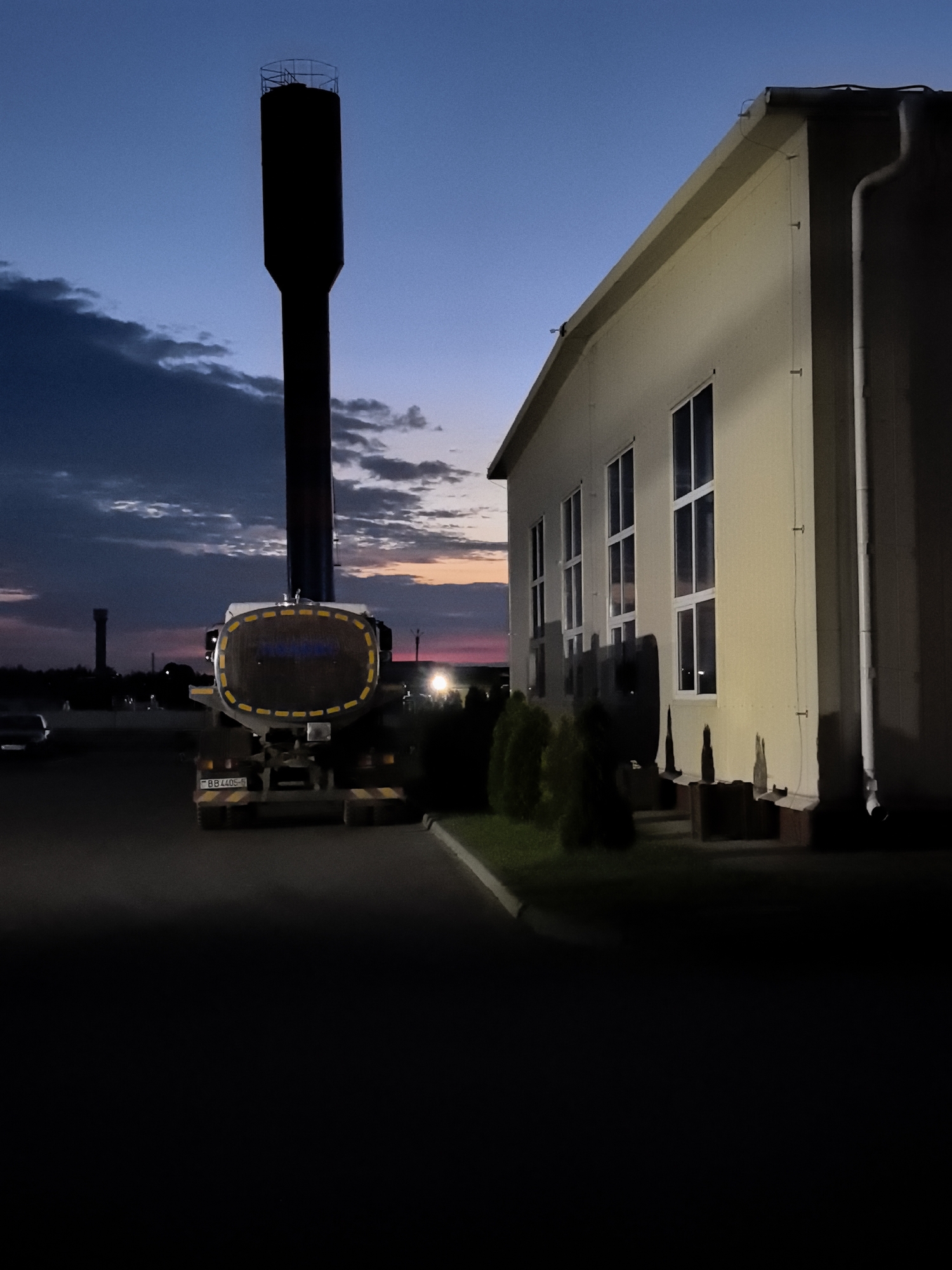 A long shot captures a water tower silhouetted against a twilight sky. The tower is tall and slender, with a wider base and a platform at the top. In the foreground, a tanker truck is parked, its trailer a large, cylindrical tank with reflective yellow markings. To the right of the tanker is a cream-colored building with multiple windows. The sky is a deep blue, transitioning to hues of pink and orange near the horizon, with scattered clouds. The scene appears to be at dusk or dawn in an industrial or semi-rural area.