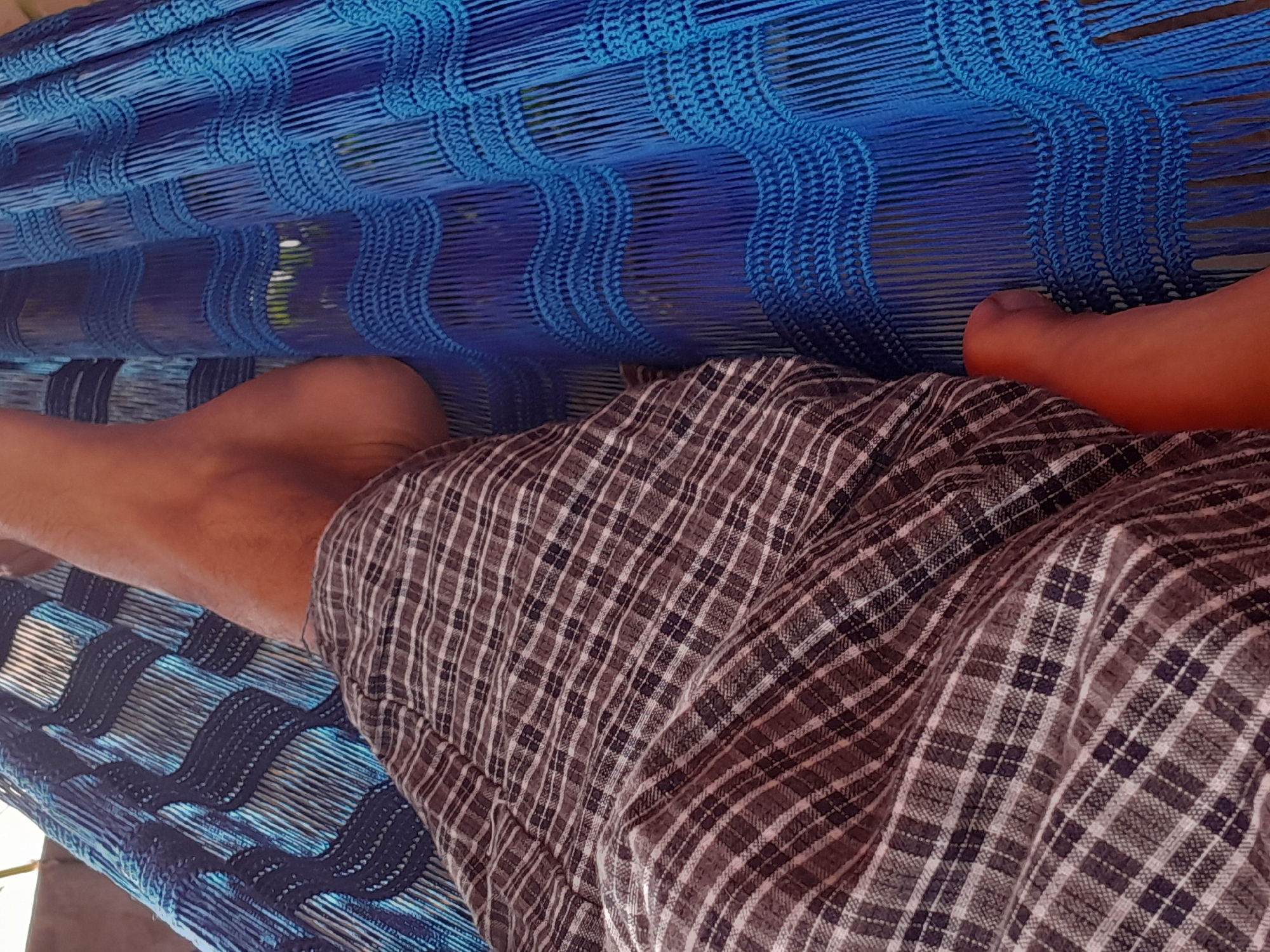 The image shows a close-up perspective of a person lying in a blue hammock. The hammock is made of woven cords and has a wave-like pattern. The person is wearing plaid shorts, and their legs are visible, along with one foot. The background is blurred but suggests an outdoor setting with some greenery visible through the hammock. The lighting indicates it is daytime. The scene evokes a sense of relaxation and leisure, typical of someone enjoying a hammock on a warm day. No text is visible in the image.