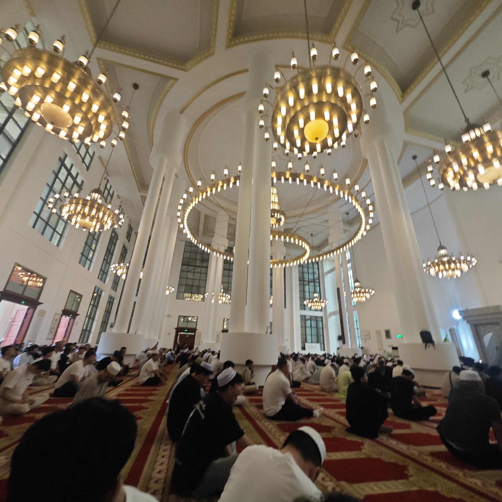 The image captures a scene inside a mosque, likely located in the Jiulongpo District, China. Numerous people are seated on a red patterned carpet, oriented towards the front of the room, suggesting they are engaged in prayer or worship. Most of the men are wearing white caps. The architecture of the mosque is characterized by high ceilings, large windows, and prominent white columns. A series of ornate chandeliers hang from the ceiling, providing illumination. The overall atmosphere appears reverent and peaceful.