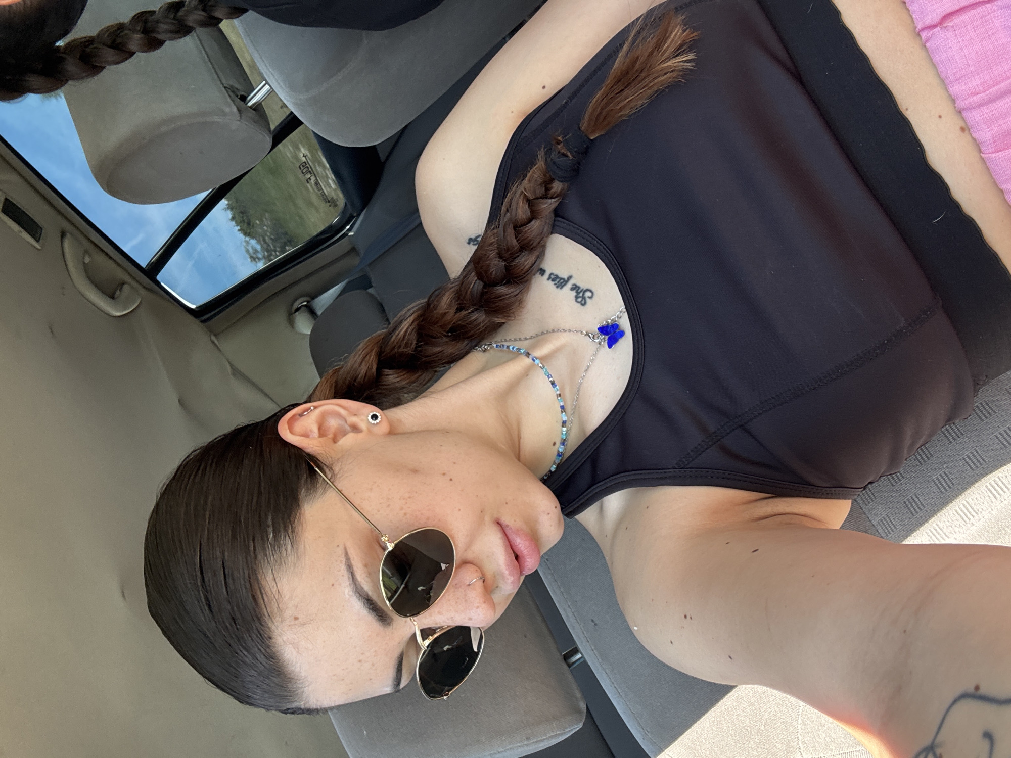 The image shows a person, likely female, in a reclined position inside a car, captured from a slightly upward angle. The individual has dark brown hair styled in a long braid that rests across their chest. They are wearing a black tank top or sports bra.
The person is wearing round, dark-tinted sunglasses. Accessories include a black stud earring and a silver hoop or bar piercing in the upper cartilage of their left ear, as well as a small silver hoop nose piercing. They have two necklaces: a multicolored beaded chain (featuring blue, green, white, and silver beads) and a silver chain with a distinct blue butterfly charm.
Several tattoos are visible: script on the upper chest/shoulder area that partially reads "She flies w", and a partial design on the lower right arm/wrist.
The setting is a car interior with gray upholstered seats and headrests. A sunroof above reveals a clear, bright blue sky with faint clouds, suggesting daytime. Through the sunroof, some greenery, likely trees, can be seen in the distance. The presence of sunlight casting shadows within the vehicle confirms bright, clear weather. On the far right, a section of pink fabric is visible. Faintly reflected in the sunroof, the word "tears" can be seen. The overall scene conveys a relaxed moment during a daytime drive in what is identified as Belopoptsi, Bulgaria.