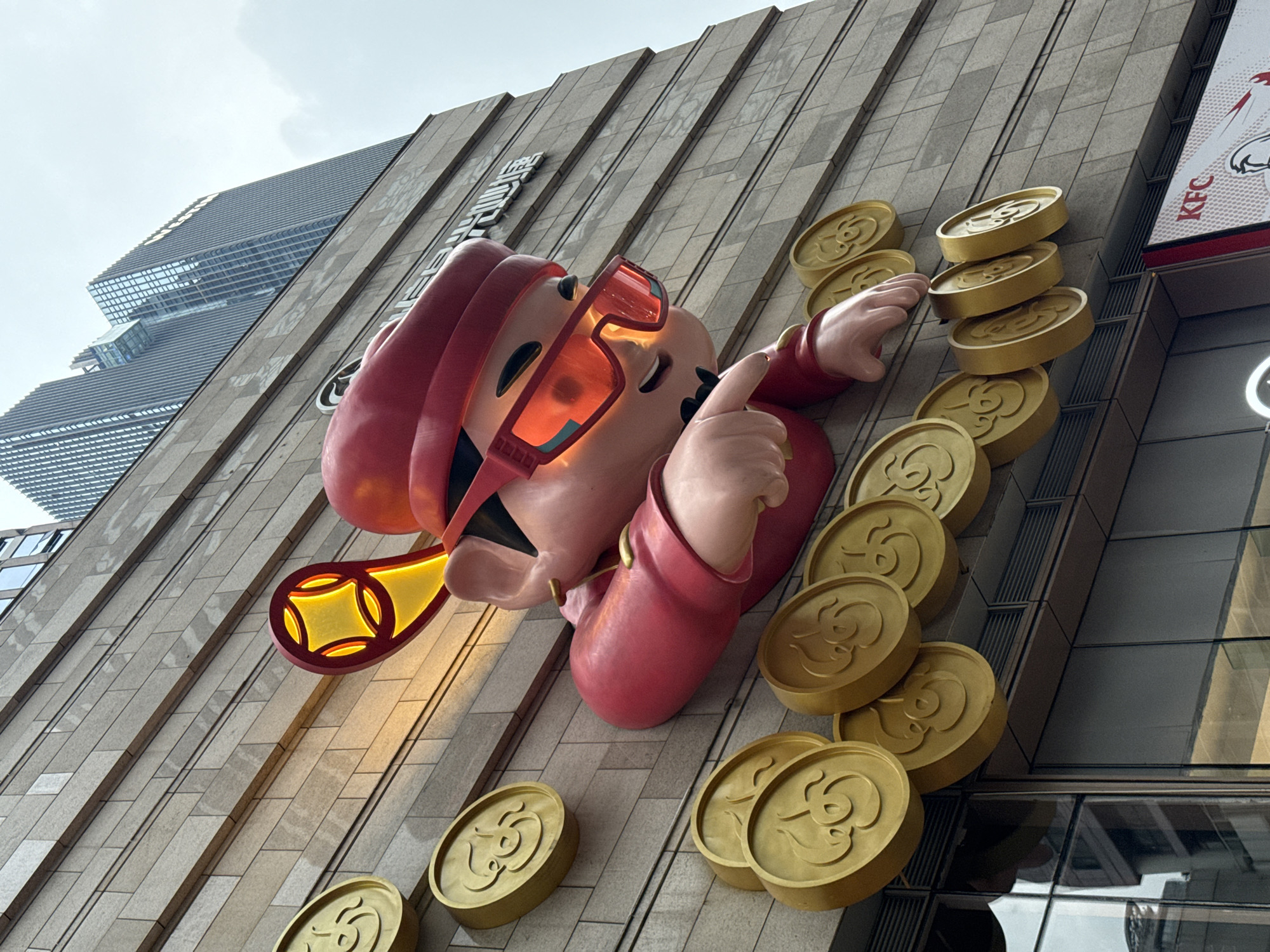 The image captures a large, cartoonish statue of a character adorned in red clothing and a beret, with oversized, bright red sunglasses. The character is situated on the facade of a modern building in Yuzhong District, China. The statue is surrounded by oversized, golden coins, each embossed with a swirling design. The character's right arm is extended, with a finger pointing downwards. Behind the statue, the building's facade is constructed of light-colored stone tiles arranged in horizontal rows. Several stories above, more modern, glass-fronted skyscrapers rise against an overcast sky. In the lower right corner, a KFC advertisement is partially visible. The perspective is low-angle, looking up at the statue and the buildings.