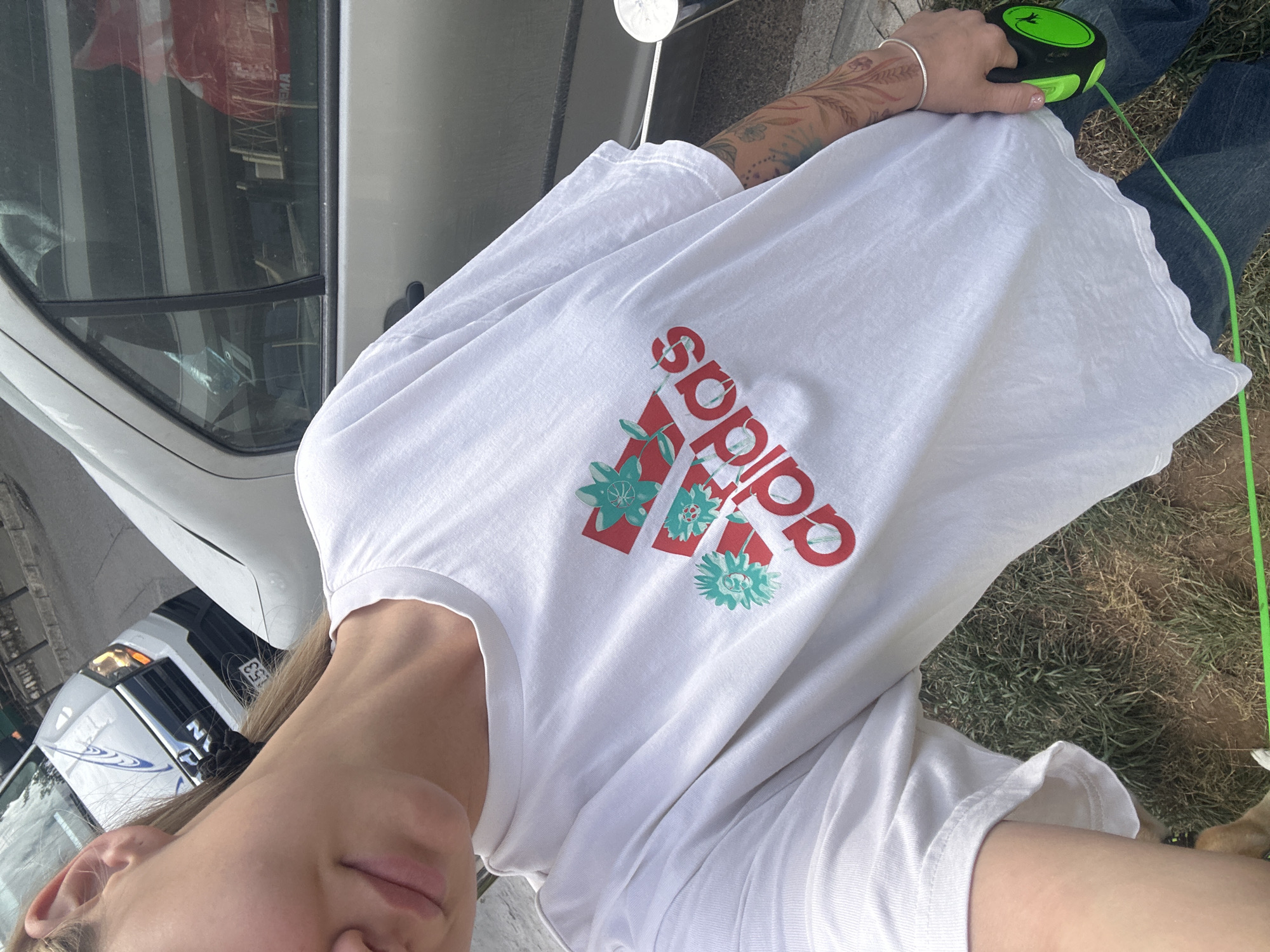 A woman is taking a selfie while standing outside. She is wearing a white Adidas T-shirt with a red logo accented with green flowers and is holding a green retractable dog leash. She is wearing jeans and a silver bracelet. Behind her is a white van and a white police car. The location appears to be near parked vehicles and grass. It cannot be determined that this is in Budva, Montenegro.