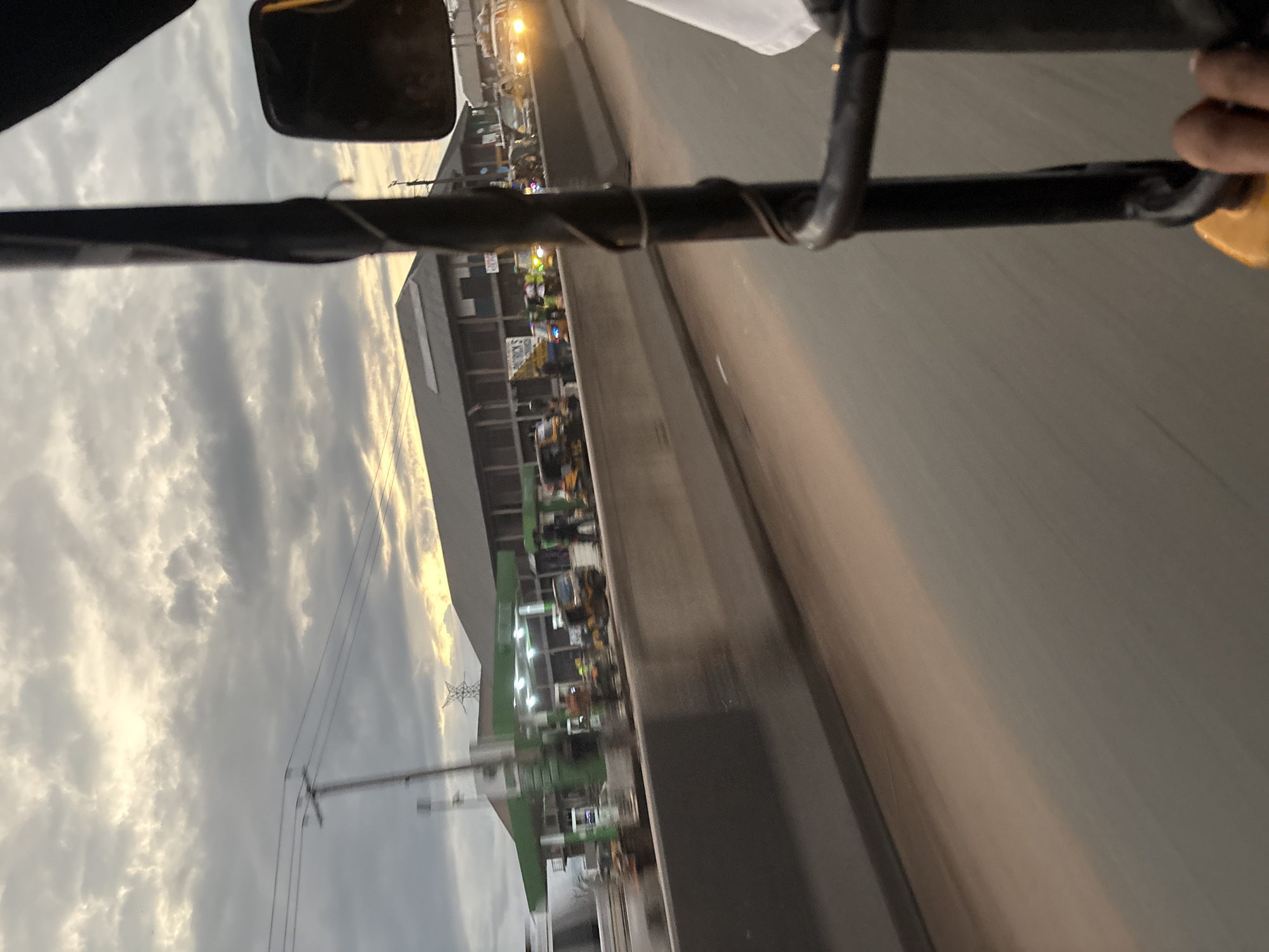 The image depicts a street scene in Onitsha, Nigeria, likely during the late afternoon or early evening, given the muted light and cloudy sky. The perspective is from the interior of a three-wheeled auto-rickshaw, with a black metal bar and a hand visible in the foreground.
In the middle ground, a concrete barrier separates the road from a row of buildings. On the other side of the barrier, several auto-rickshaws are parked or moving. A prominent green-roofed structure, appearing to be a gas station, is visible with pumps and lights. People are seen near the gas station and the auto-rickshaws. Further down the road, other vehicles, including cars with illuminated headlights, are present. The sky is filled with textured clouds, with some lighter areas suggesting the presence of the sun or a break in the cloud cover. The overall impression is of a busy urban environment.