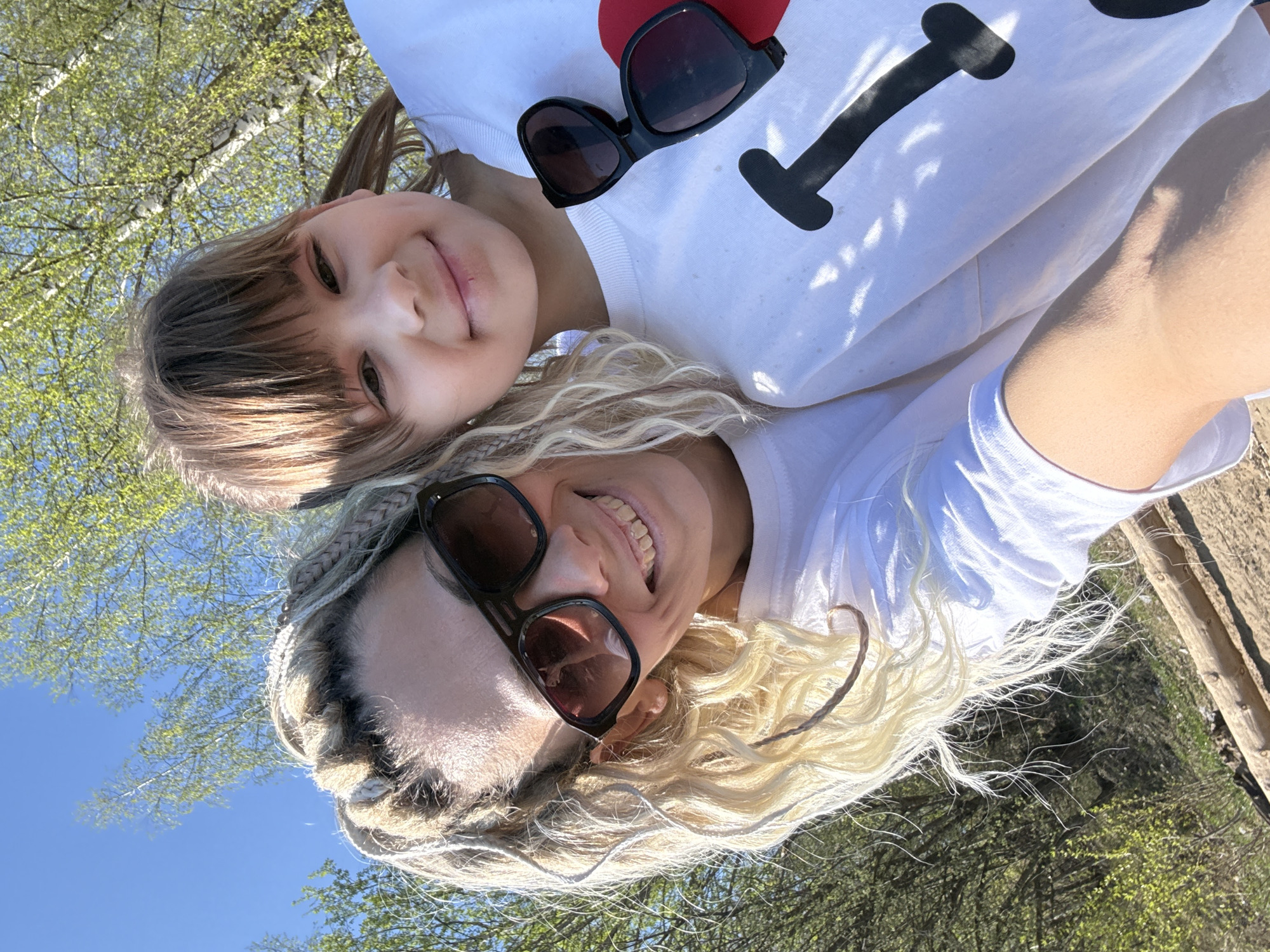Two individuals, presumably a child and an adult, are captured in a selfie-style photograph outdoors. The child is positioned slightly above and behind the adult. The child has light brown hair with bangs and is wearing a white shirt with a large black "I" and a red heart graphic. A pair of sunglasses rests on the shirt near the neckline. The adult has long, blonde hair in braids and is wearing sunglasses with brown lenses and a white t-shirt. Both individuals are smiling at the camera.
The background suggests an outdoor setting with a blue sky and trees with foliage visible. The location is identified as Rusanovo, Russia. Part of a wooden structure is also visible in the background.
