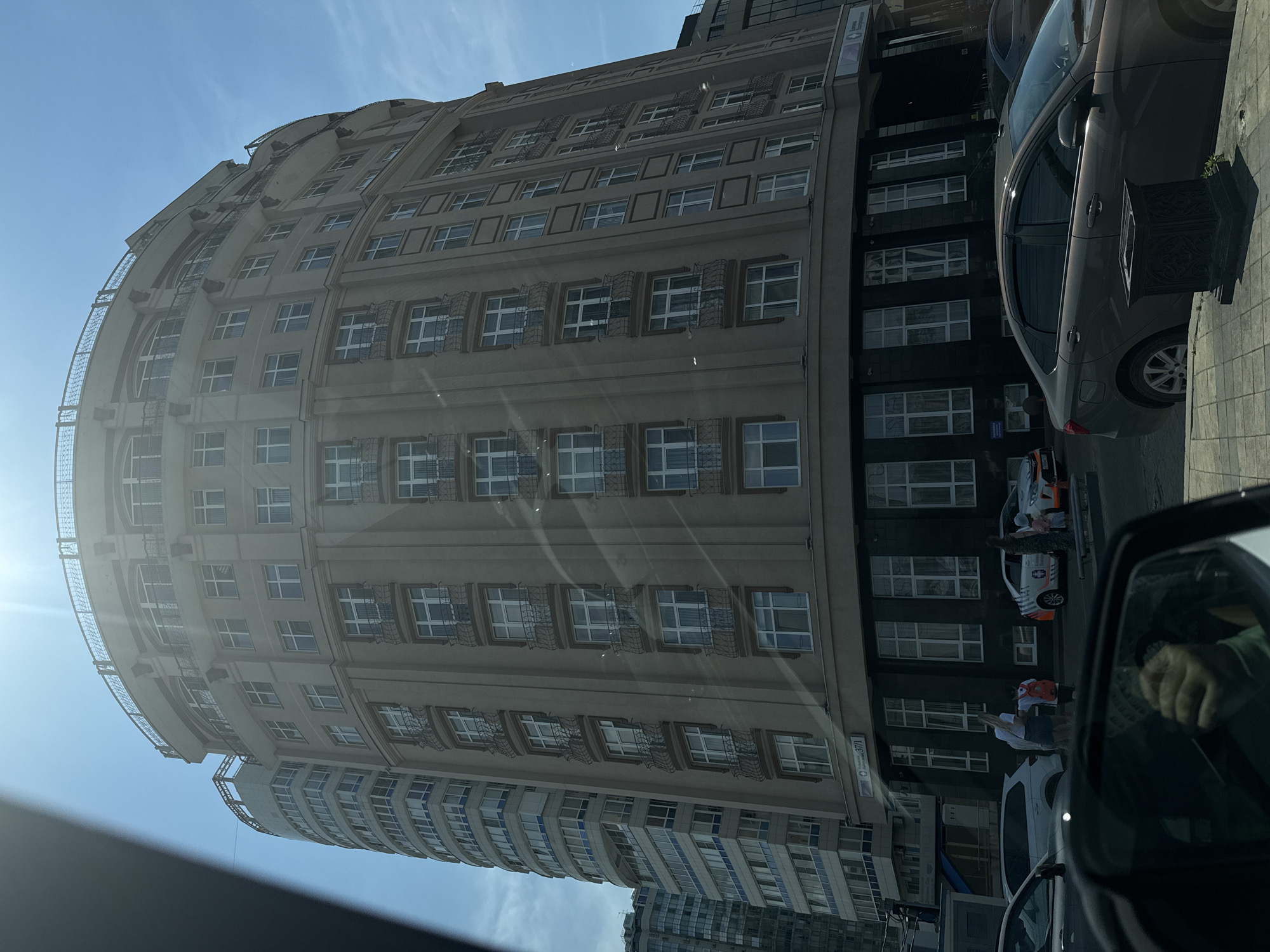 This is a photograph of a modern, curved building with a light-colored facade and numerous windows. The building appears to be residential or commercial, with a mix of window styles, some featuring decorative ironwork. The architecture suggests a contemporary design.
In front of the building, a dark-colored car is parked on a sidewalk. Next to the car, there is a large, black planter. Further down the street, another car with an orange and white design is visible, possibly a service vehicle, with people near it. The presence of cars and a paved sidewalk indicates an urban setting.
The scene is bathed in sunlight, casting shadows and creating a lens flare effect in the upper left portion of the image, suggesting it is a sunny day, likely mid-morning or early afternoon. The sky is clear and blue.
There are faint reflections on the windows of the building and a slight glare across the image, possibly from the camera lens or a protective screen. The perspective of the shot is from a low angle, possibly from inside another vehicle looking out. The image is oriented vertically, with the building curving upwards.
Visible text is limited to a small sign on the building that appears to read "37/1". The overall impression is a snapshot of urban street life on a bright, clear day.