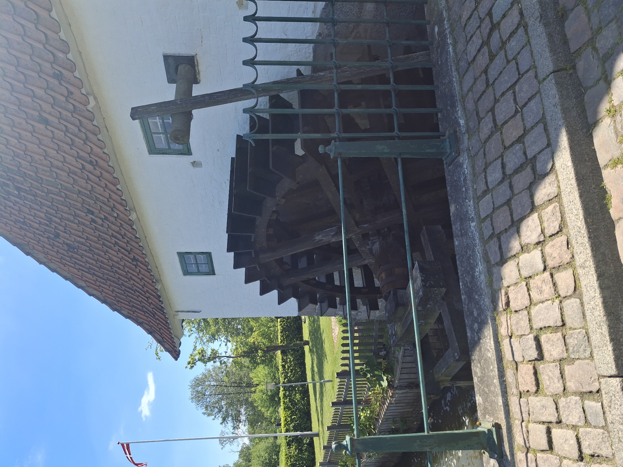 The image depicts a watermill, possibly historical, adjacent to a paved area. The mill is characterized by a large wooden water wheel, partially obscured by a green metal fence. The mill building itself has white walls and a red tile roof. Two small, square windows with green frames are visible. In the background, a green lawn and trees are present, suggesting a park or garden setting. A Danish flag is present on a flagpole in the background. The scene is bathed in daylight. The location is Unknown, Unknown.