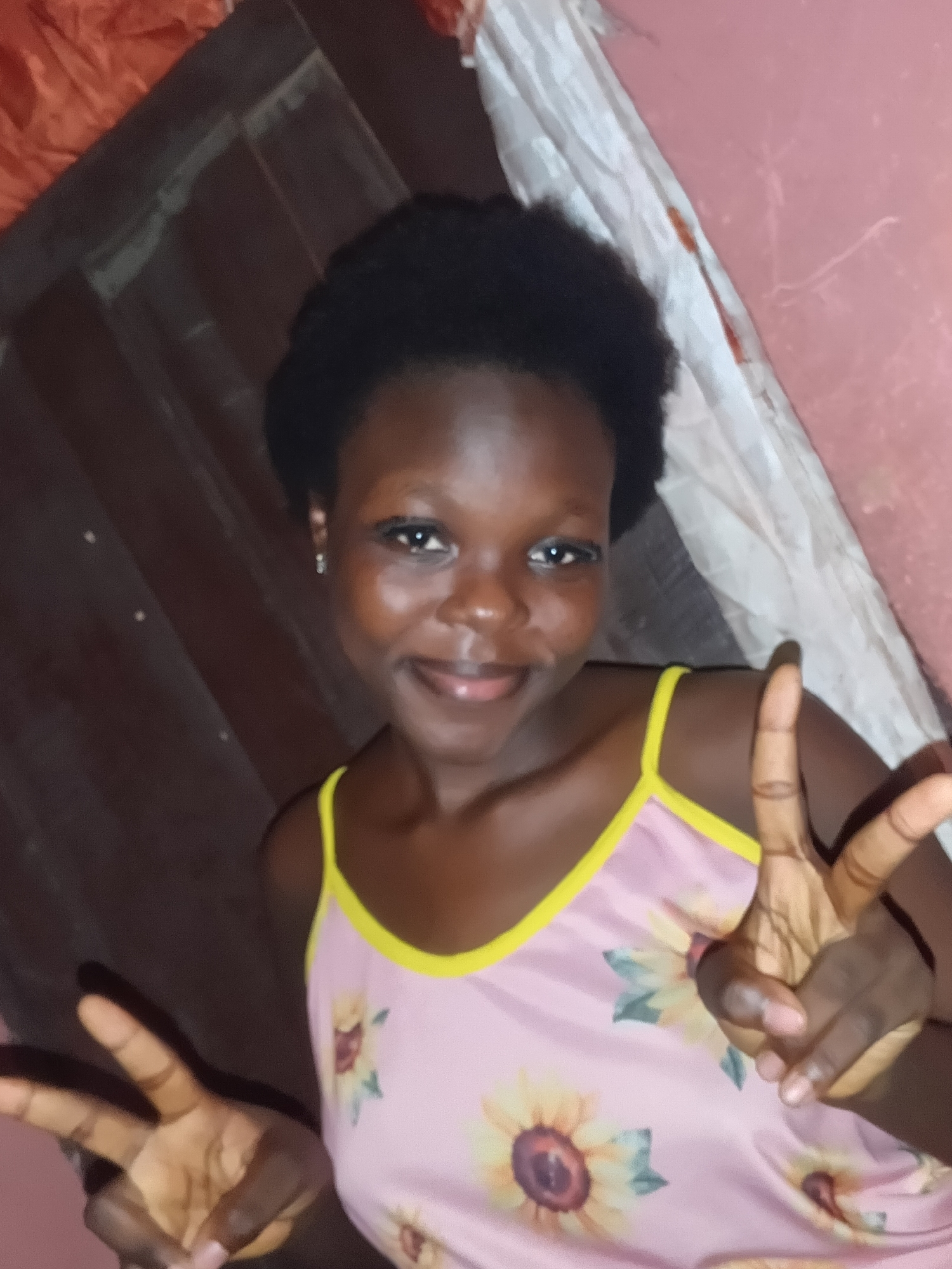 A young, dark-skinned female is depicted in a chest-up portrait. She possesses short, dark, curly hair and a subtle smile. She is wearing a light pink camisole with yellow spaghetti straps and yellow trim, adorned with a sunflower print. Both of her hands are raised, each forming a "peace sign" (V-sign) with the index and middle fingers extended.
The background to her left consists of a dark brown, possibly corrugated or paneled surface, with some faint horizontal lines and reflective dots. An orange crumpled fabric is partially visible in the upper-left corner. To her right, the background is a light pink wall. A piece of white fabric, possibly crumpled or draped, runs diagonally behind her right shoulder, exhibiting a reddish-brown stain or mark. The lighting appears dim and uneven. The image provides no discernible information to confirm the location as the city of Onitsha, Nigeria.