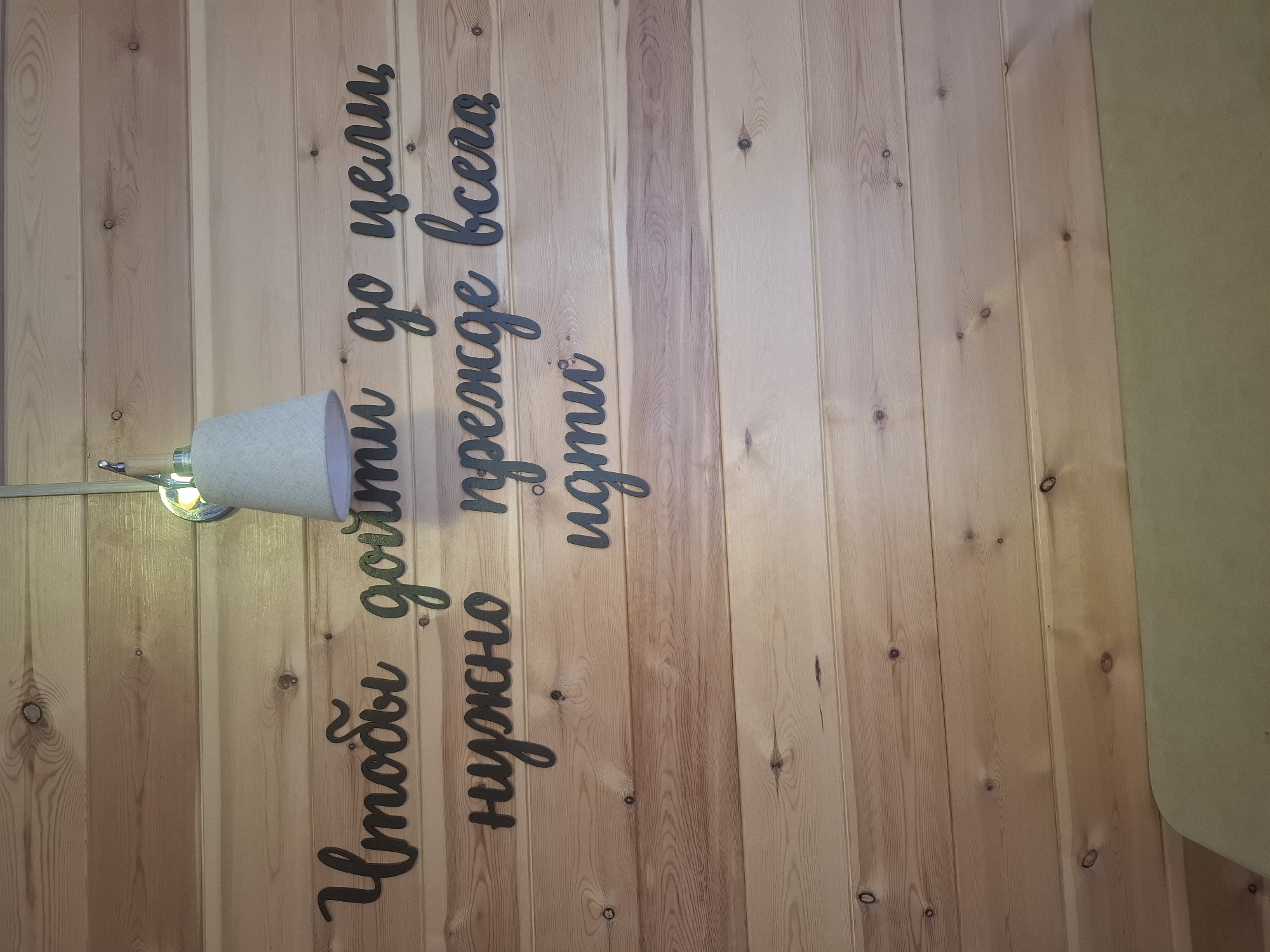 The image shows a wooden wall with black, cursive text cutouts. The text reads "Чтобы дойти до цели нужно прежде всего идти". This translates to "To reach the goal, one must first walk". Above the text, a light fixture with a conical shade is mounted on the wall. The overall setting appears to be an interior space with natural wood paneling. The location in the city of Sukhoy Ruchey, Russia, is not discernible from the image itself. There are no people or discernible actions depicted. The scene is static and focuses on the wall decoration.