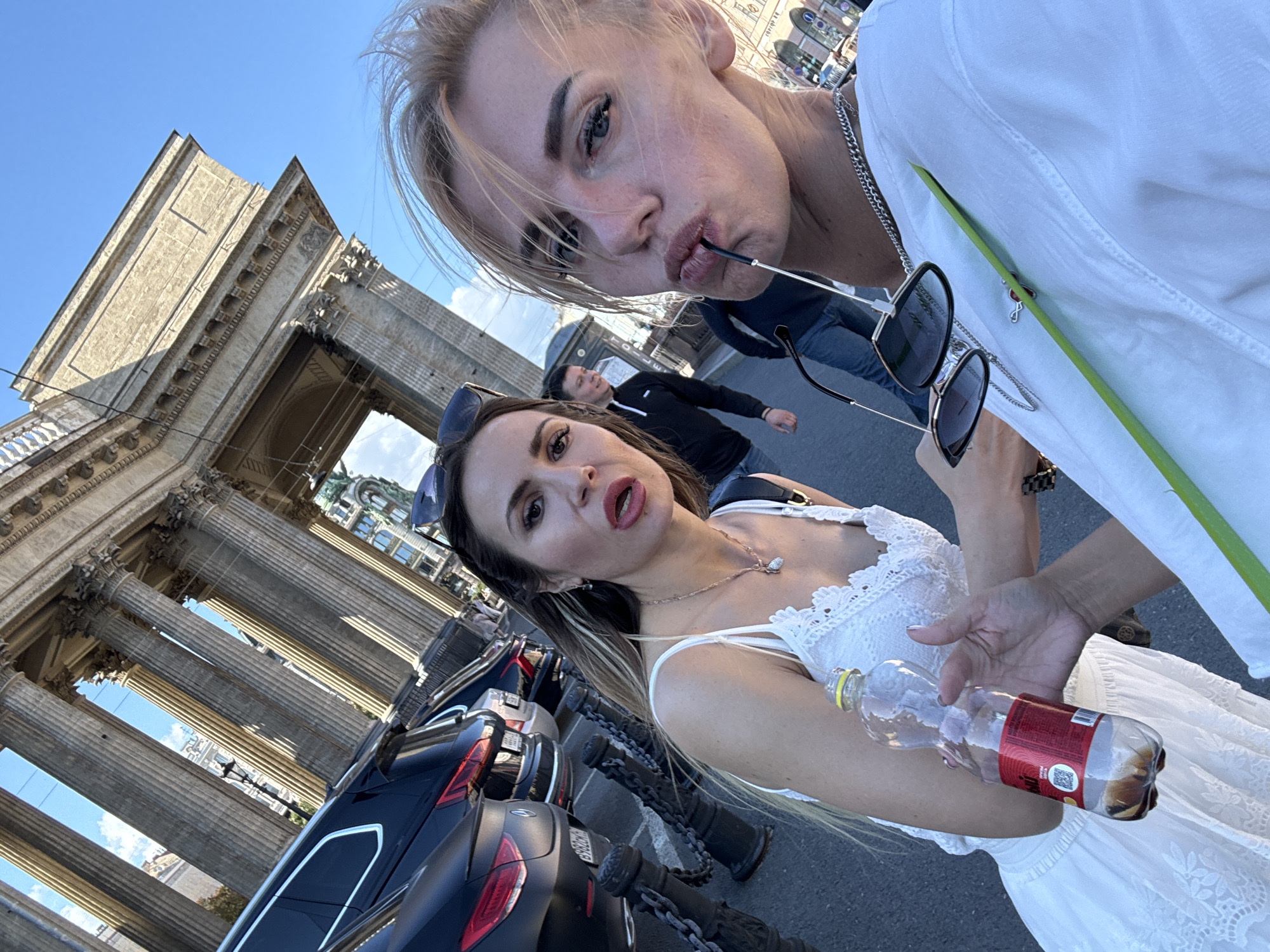 Two women are taking a selfie in front of the Kazan Cathedral in Saint Petersburg, Russia. The woman on the left is holding a bottle of water. The woman on the right is holding a pair of sunglasses. In the background, several cars are parked and two men are walking. The sky is clear and blue.