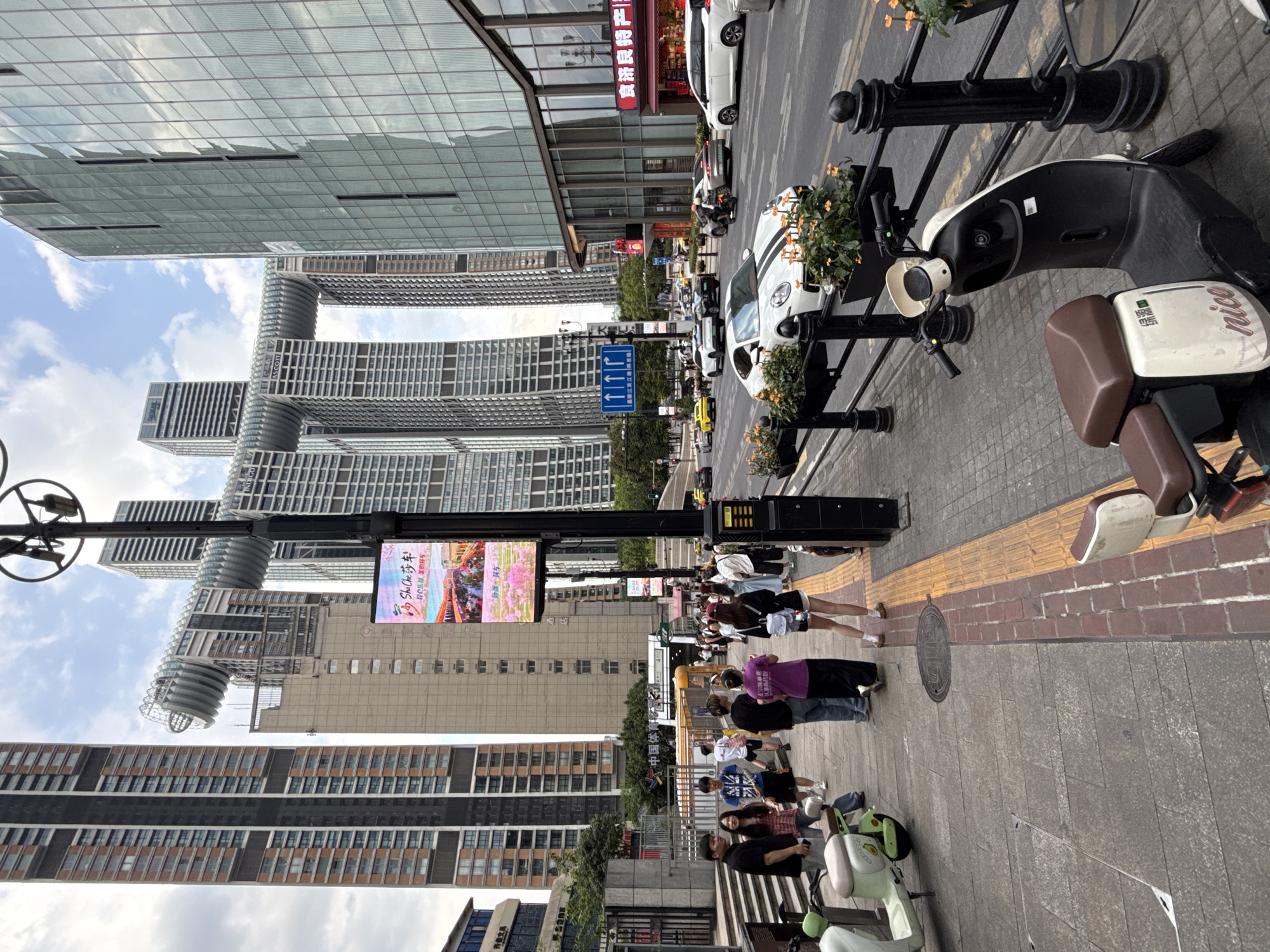 The image captures a street scene in Yuzhong District, China, characterized by a cluster of modern skyscrapers, including the distinctive Raffles City Chongqing with its horizontal connecting structure. In the foreground, a sidewalk is populated by pedestrians, some standing and others walking. Several electric scooters are parked along the sidewalk.
The sidewalk itself is paved with stone slabs and features a tactile paving strip for visually impaired individuals, demarcated by yellow and red brick sections. A black lamppost with signage and a digital billboard displaying a colorful advertisement stands prominently near the center of the frame.
Beyond the sidewalk, a street is visible with moderate traffic, including cars and a motorcycle. Cars, such as a white Porsche with racing stripes and a white SUV, are present. The buildings lining the street are modern and architectural, with glass facades reflecting the cloudy sky.
In the mid-ground, a subway entrance is visible, indicated by a yellow barrier and signage. People are seen entering and exiting the area around the entrance. The overall environment suggests an urban setting with a mix of commercial and residential structures, bustling with pedestrian and vehicular activity under a partly cloudy sky.
