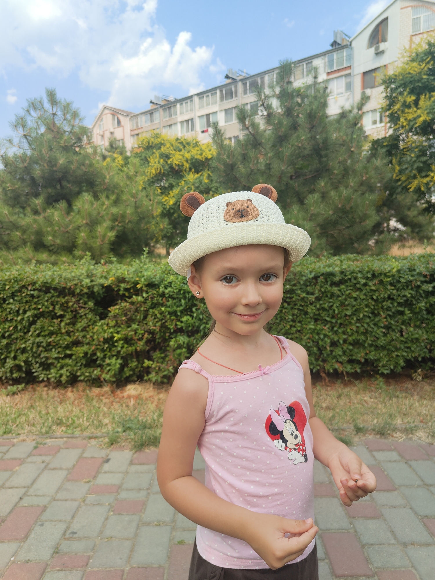A young girl with light brown hair and brown eyes smiles at the camera. She is wearing a white bucket hat with bear ears and a brown bear face applique. The hat also has a decorative string around it. She is also wearing a pink tank top with white polka dots and a graphic of Minnie Mouse within a red heart. Her skin tone is fair, and she has a small, delicate earring in her left ear.
The girl is standing outdoors, with a paved walkway of reddish-brown and gray rectangular stones in the foreground. Behind her is a neatly trimmed green hedge, and further back are pine trees and a building with multiple windows. The sky above is blue with scattered white clouds, suggesting a pleasant day. The lighting indicates it is daytime, likely late morning or afternoon, with the sun casting soft shadows. The girl's expression is happy and relaxed, and she is holding something small in her hands. The overall scene suggests a casual outdoor setting, possibly a park or residential area.
There is no visible text in the image. The location context provided is Chornomorsk, Ukraine.