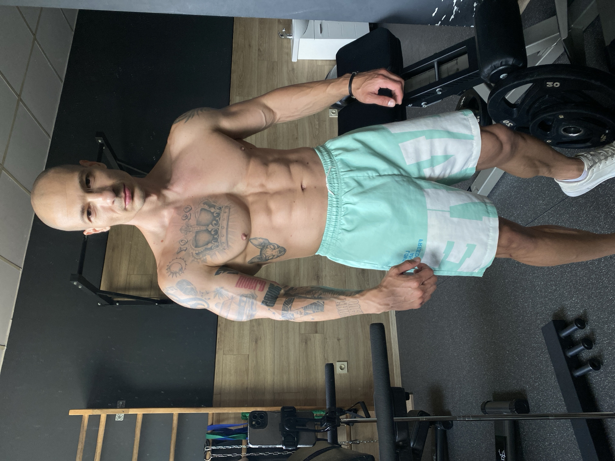 A shirtless man with a shaved head and numerous tattoos is standing in what appears to be a home gym. He is wearing light blue and white athletic shorts and white sneakers. The tattoos cover his arms, chest, and upper torso, featuring designs such as a crown, symbols, and writing. He is looking directly at the camera. The gym contains weightlifting equipment, including a leg extension machine with weight plates, a barbell, and a pull-up bar attached to a wall. The floor has both wood flooring and a dark, possibly rubberized, workout surface. The location cannot be verified as Pleven, Bulgaria, from this image alone.