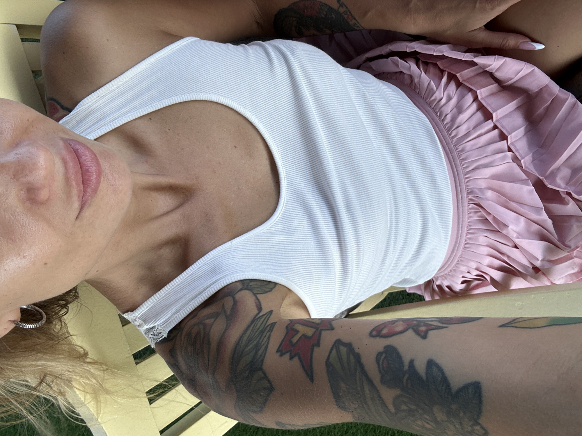 A close-up, high-angle selfie depicts an individual seated outdoors on a light yellow wooden slatted bench in Phu Thuy Ward, Vietnam. The subject, a female, has blonde hair partially visible on the left side of the frame, and wears a silver-toned, ridged hoop earring in her left ear. Her lips are light pink. She is dressed in a white ribbed tank top with a lace detail visible on the left strap, and a light pink pleated skirt. The left arm and shoulder are extensively covered with tattoos, including floral designs, abstract shapes, and a red and yellow explosion-like motif with a "Y" inside. A tattoo is also partially visible on the upper right arm. The right hand rests on the skirt, displaying nails with white tips. A green, grass-like surface is visible in the background.