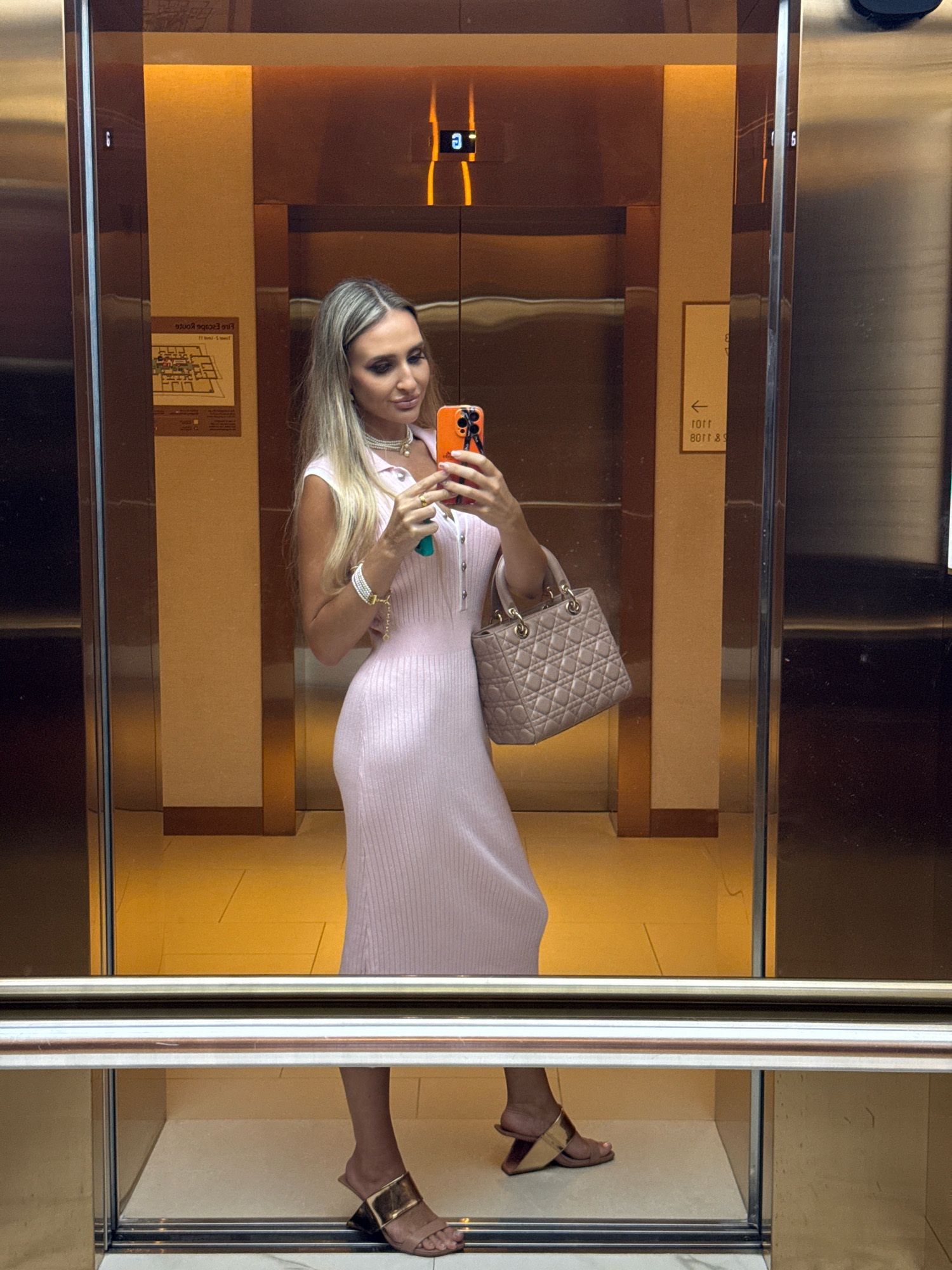 In Dubai, United Arab Emirates, a woman stands in an elevator, taking a selfie in a mirrored wall. She is wearing a sleeveless pink ribbed dress, a pearl necklace, and stacked bracelets. She holds an orange smartphone in her right hand and a light beige quilted handbag with a gold chain strap in her left hand. She is also wearing stylish open-toe mules with a gold geometric heel. The elevator's interior is lined with polished stainless steel and bronze-toned panels. The floor is a light cream color, and the ceiling features warm, indirect lighting. Reflecting in the mirrored walls are elevator control panels and signage.