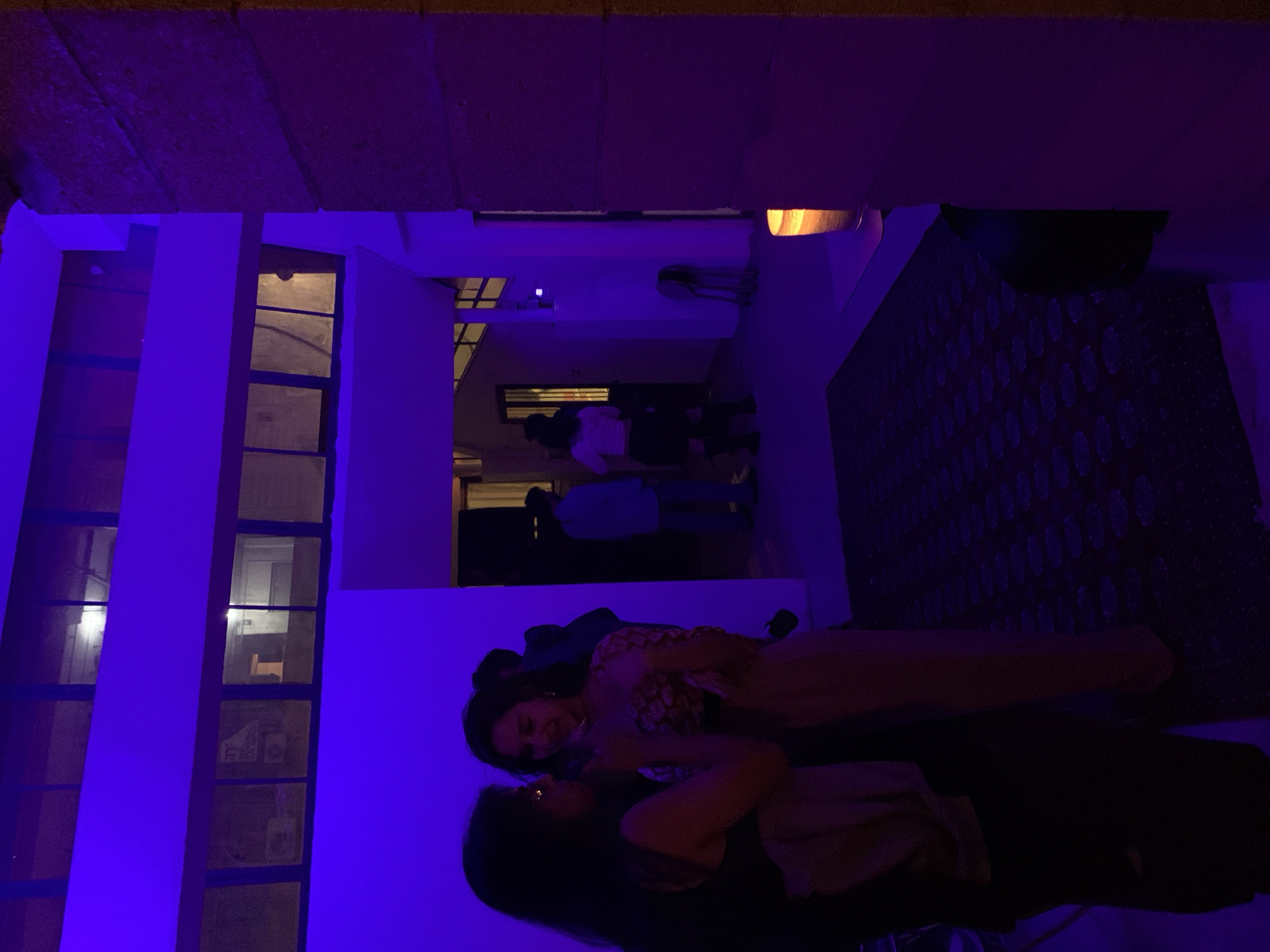 A vertically oriented photograph shows an outdoor nighttime scene in what appears to be an urban setting in Tel-Aviv, Israel. The lighting is primarily purple and dark.
In the foreground, two women are lying on a surface, possibly a bed or a lounge area. One woman is wearing a patterned top and pants, while the other is wearing a top or shirt. Their faces are visible and they appear to be interacting with each other.
In the background, there is an open-air courtyard or walkway with other individuals walking. The architecture is modern, featuring rectangular windows and walls, and appears to be illuminated with the same purple lighting. Above is visible a building. There is some art with circles on the wall.