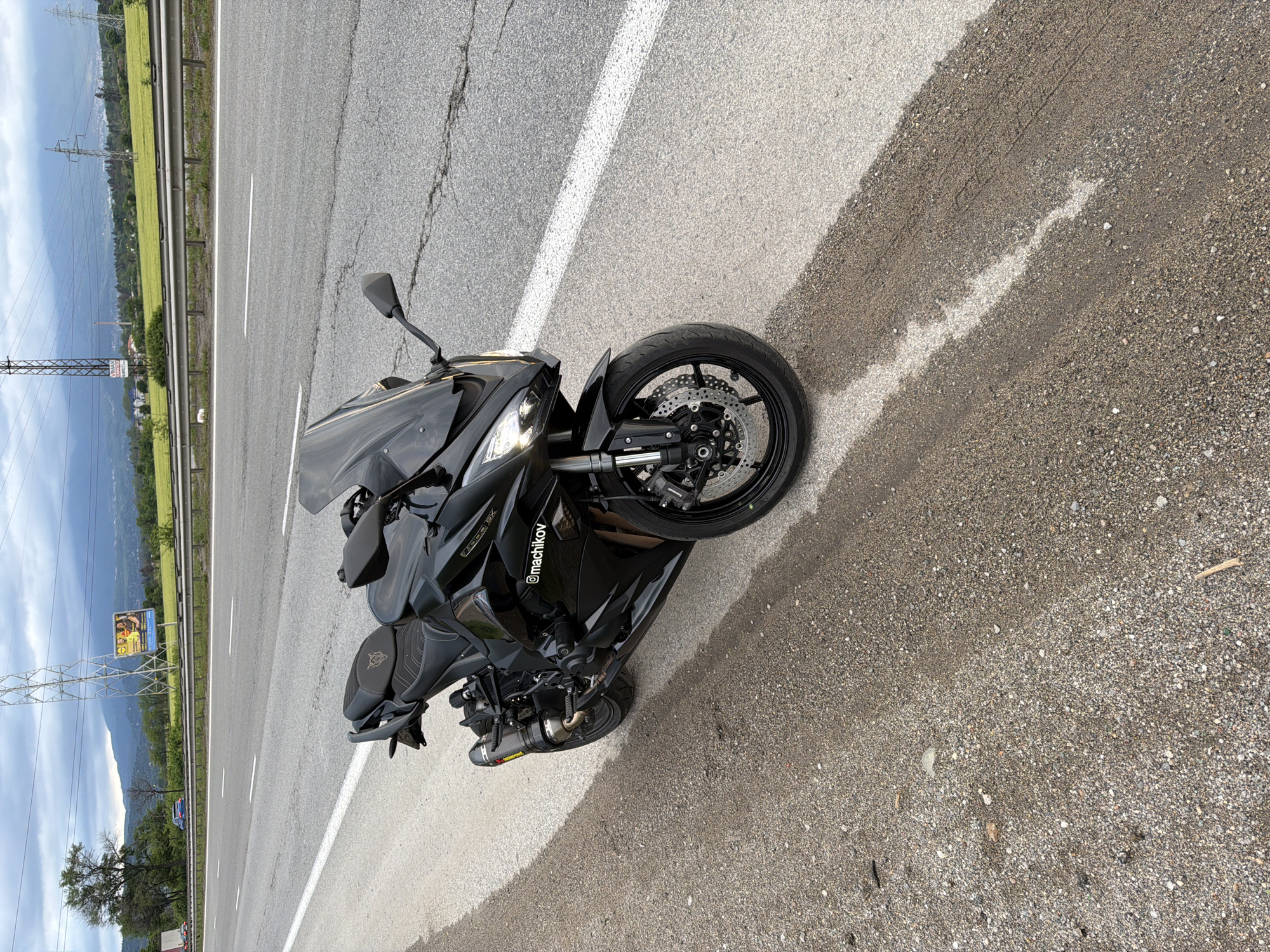 The image shows a black motorcycle parked on the shoulder of a road. The road is made of asphalt and has a white lane marker. The shoulder of the road is covered in gravel. In the background, there are power lines, fields, and mountains. A billboard is visible along the road. The motorcycle has an aftermarket Akrapovic exhaust and an inscription with the handle "@machikov". The photograph was taken in the city of Kazichene, Bulgaria.