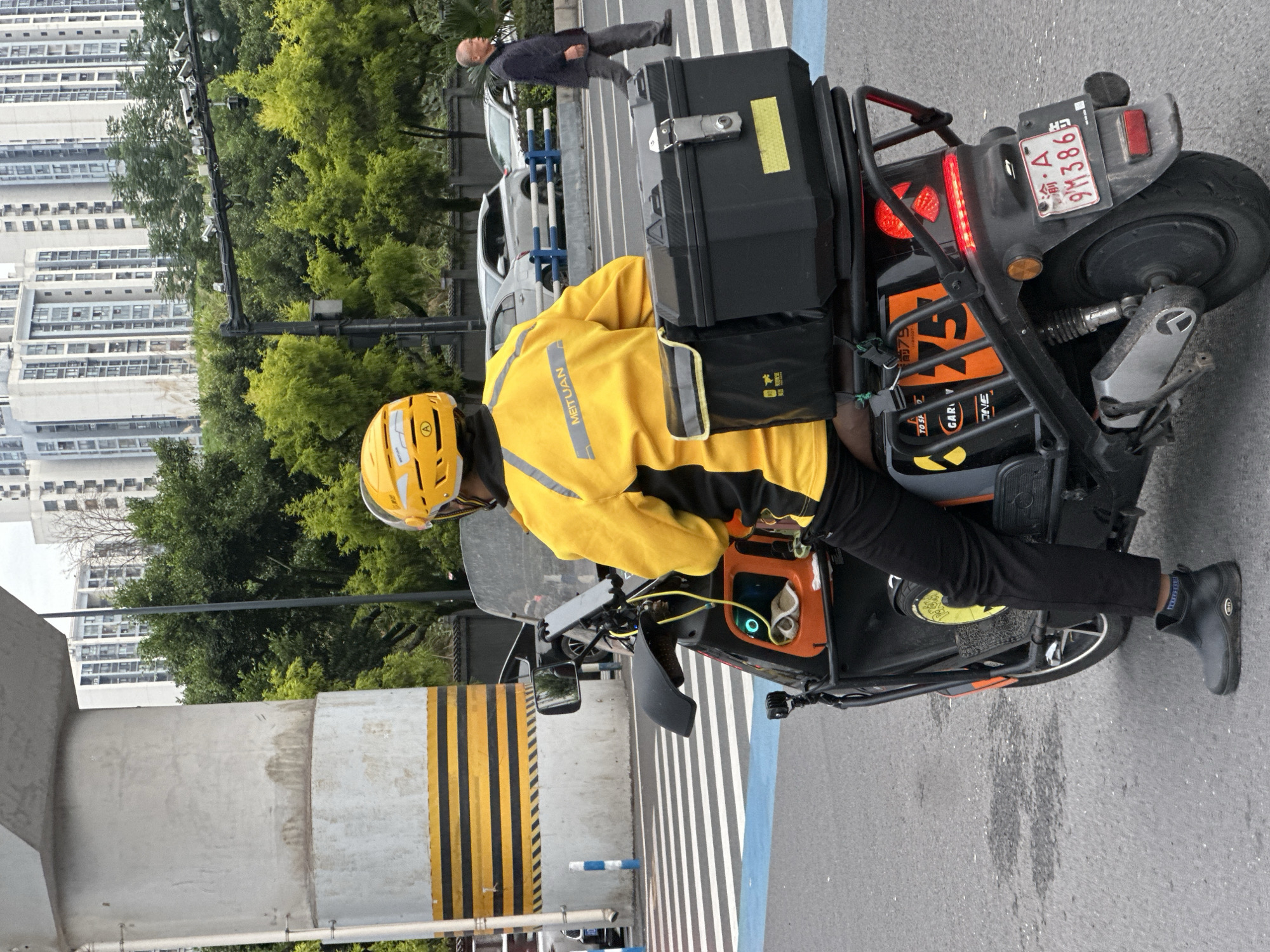 An aerial view from behind shows a person wearing a bright yellow jacket and helmet, seated on a black and orange electric scooter. The scooter has a large black storage box mounted on the rear, with a smaller black bag attached to its left. The rider's jacket has the "MEITUAN" logo. The scooter is positioned on an asphalt road, with a pedestrian crossing clearly marked with white stripes and blue accents.
In the background, a large, grey concrete pillar with black and yellow hazard stripes stands prominently on the left. Beyond the pillar, lush green trees are visible, interspersed with modern, tall residential buildings. A man in a dark jacket and grey pants is walking across the pedestrian crossing in the middle ground. Several cars are parked or moving on the road behind the pedestrian crossing. The scene appears to be captured in an urban environment, likely in the city of Chongqing High-tech Zone, China, suggested by the context. The lighting indicates an overcast day.