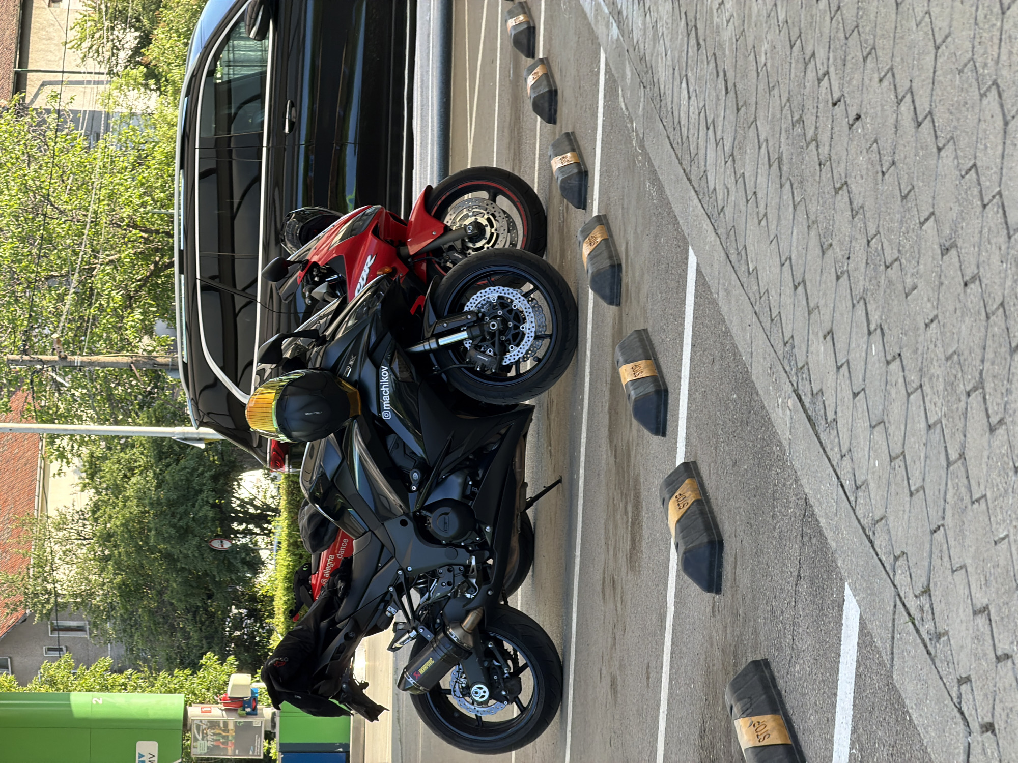 The image depicts a parking area with two motorcycles parked next to a black car. The motorcycles, one red and one black, are adjacent to a row of black parking stops. A gas station with a green MV sign is visible in the background. The location appears to be in an urban setting, evidenced by the architecture and trees in the background. Based on the gas station brand, it's likely the location is in Sofia, Bulgaria.