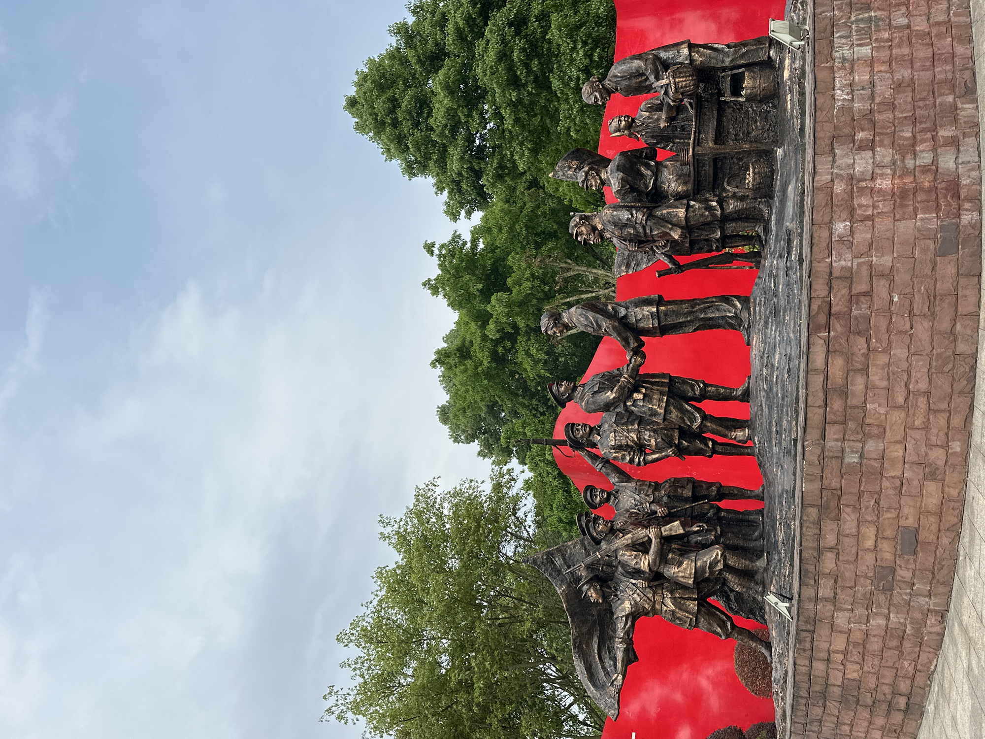 The image depicts a bronze sculptural monument against a background of green trees and a partly cloudy sky. The monument features a series of figures in a row, set against a vibrant red backdrop. The figures are depicted in what appears to be military or revolutionary attire. They are positioned on a base constructed of brickwork. The monument is located in Liangjiang, China.