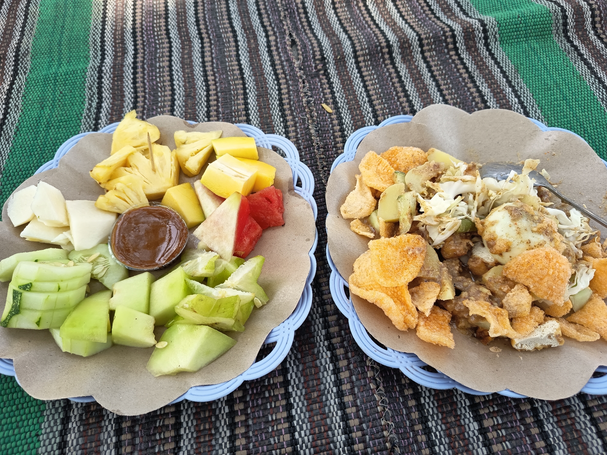 Two blue plastic plates, each with a brown paper liner, are placed on a striped mat. The plate on the left contains a selection of cut fruits, including pineapple, watermelon, melon, and possibly guava, served with a small cup of dark dipping sauce. The plate on the right contains a dish composed of various ingredients including fried crackers, chopped vegetables, and what appears to be hard-boiled egg, all coated in a sauce. A metal spoon rests on the edge of the right plate. The setting suggests a casual outdoor meal, possibly in Lalung, Indonesia.