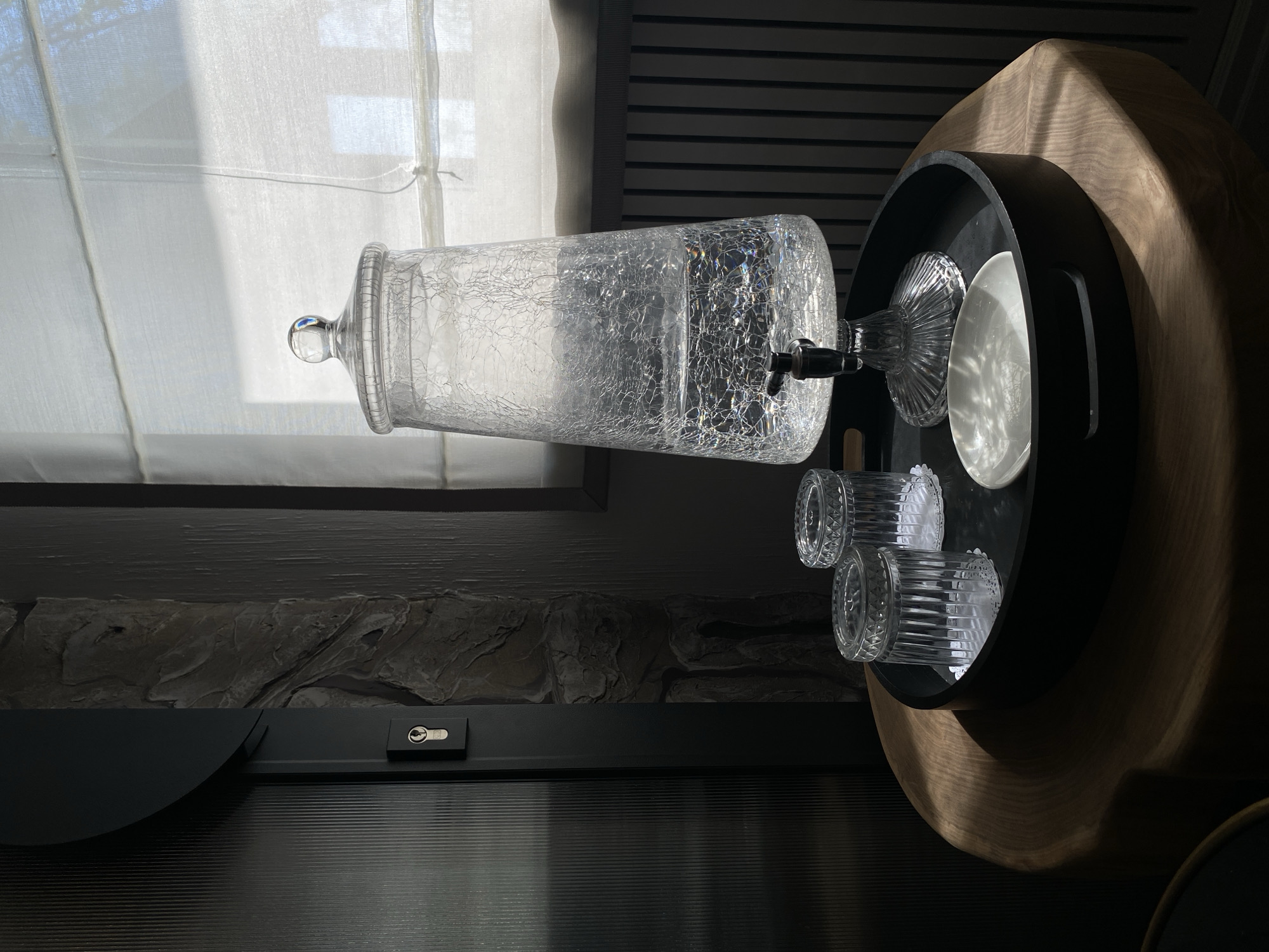 The image shows a glass water dispenser filled with water, a water glass, two drinking glasses, and a white plate, all arranged on a black tray. The tray is placed on a light-colored wooden side table with a curved edge. Behind the side table, there is a window covered with a white fabric blind, and a dark-colored wall with a stone texture. The scene is indoors and well-lit. The location is not discernible from the image and there is no indication that it is in the city of Votkinsk, Russia.