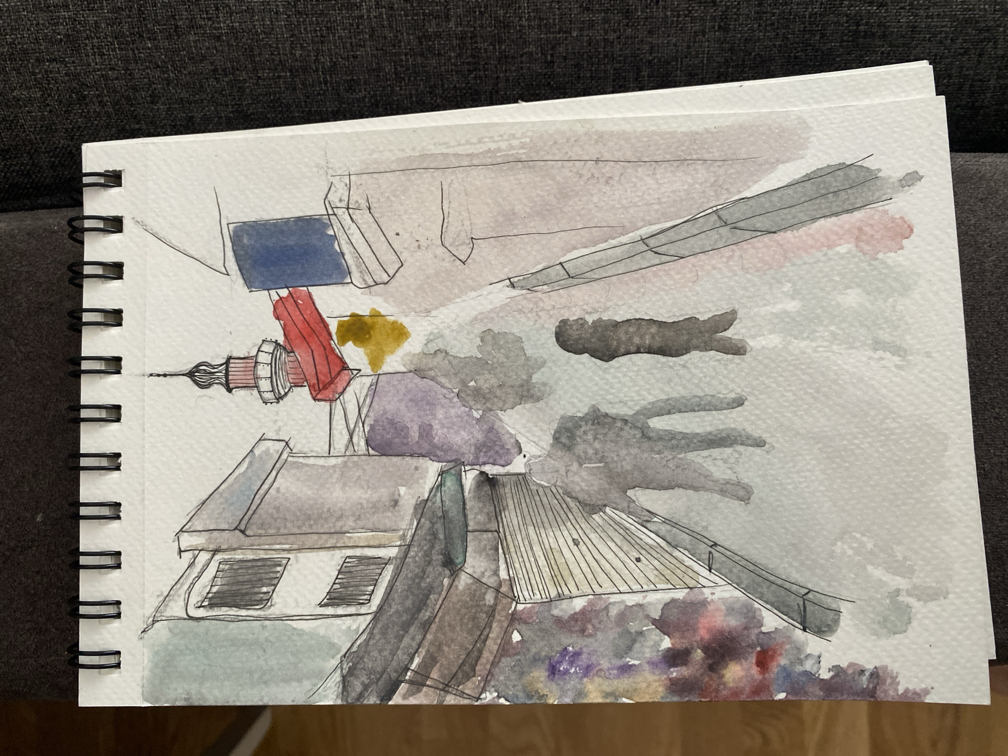 A watercolor sketch in a spiral-bound notebook depicts an urban scene with buildings and a tower. The tower is thin and tall, with red and white sections. Buildings of various heights and shapes surround the tower. Some appear to have flat roofs and windows. Trees are represented with purple and grey watercolor washes. The sky is a light grey watercolor wash. The location is in the city of Unknown, Unknown.