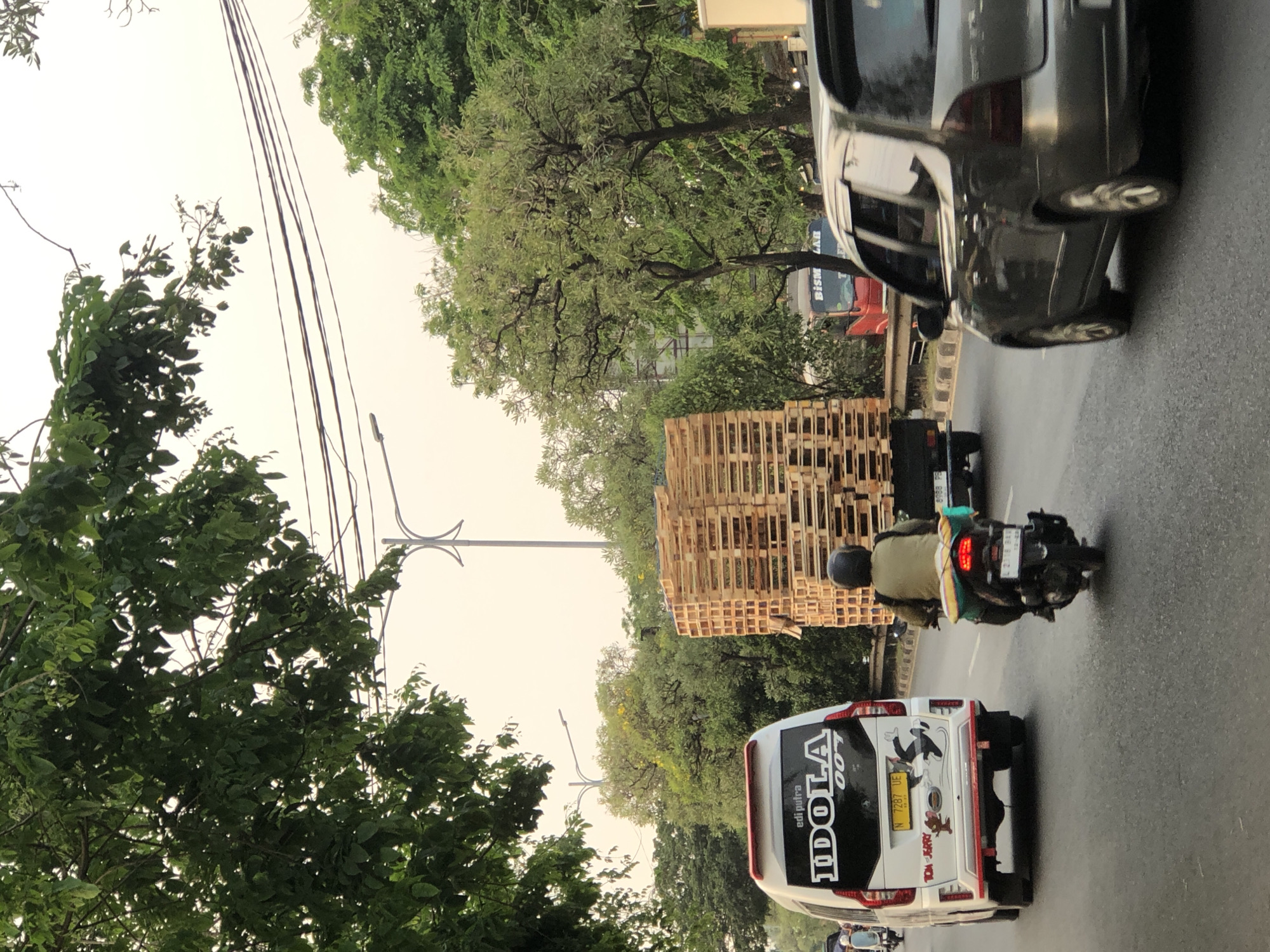The image depicts a street scene in Kludan, Indonesia, characterized by a moderately busy road with several vehicles. In the foreground, a gray car is visible on the right, with its rear section showing. To the left, a white van with the words "edi putra IDOLA 007" and cartoon characters Tom and Jerry is prominent. A motorcycle with a rider wearing a helmet and a green jacket is positioned between the van and a truck laden with wooden pallets. The truck, a dark color, is the largest object in the frame, its cargo of stacked wooden pallets dominating the center of the image. In the background, various trees line the street, interspersed with utility poles and overhead power lines. The sky is overcast, suggesting a muted natural light. The overall impression is that of a typical daytime traffic scene in an urban or suburban environment.