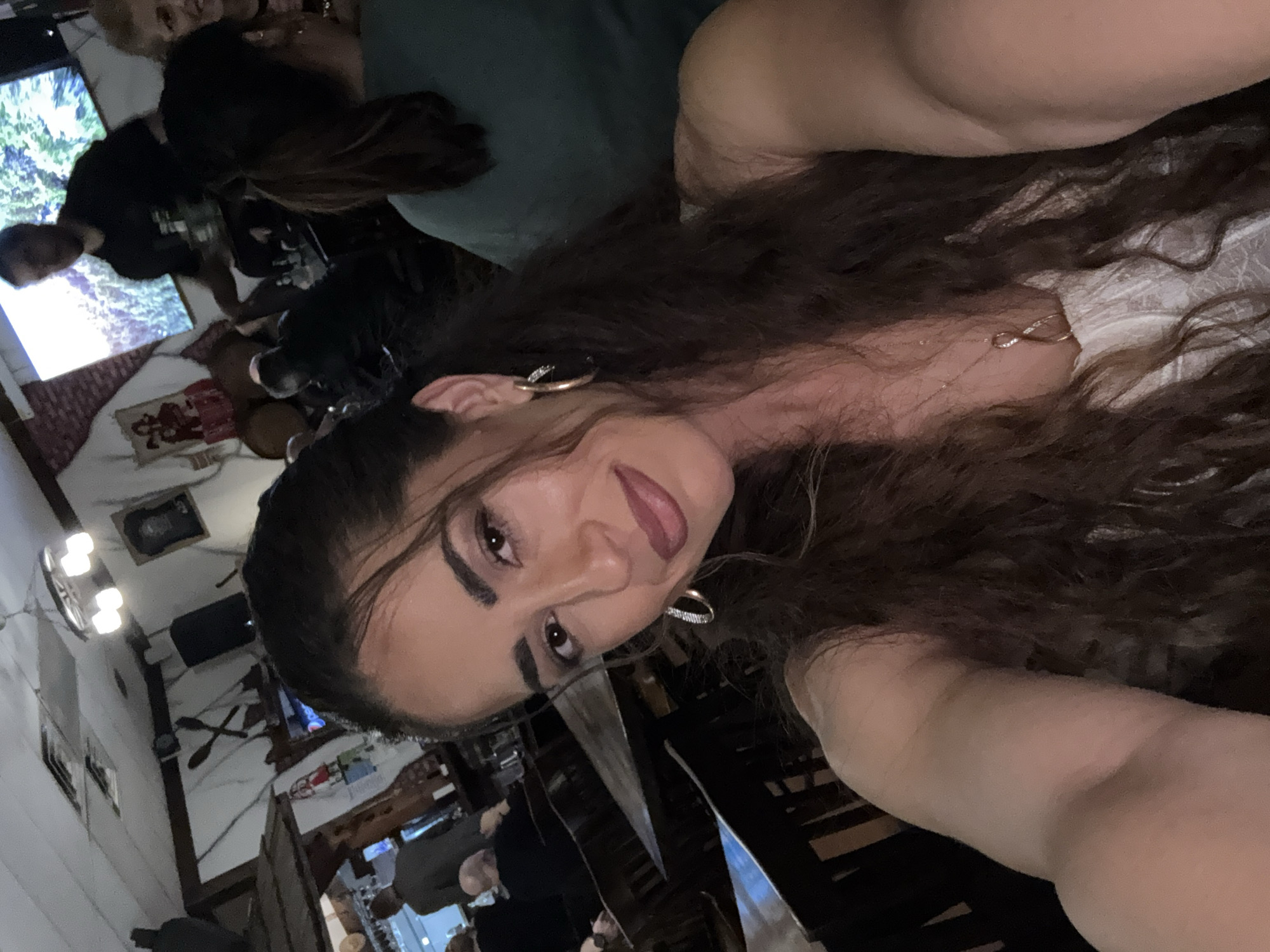 A selfie taken in an indoor dining area in Des Plaines, Illinois. The person taking the photo has long, dark, curly hair, and is wearing gold hoop earrings and a delicate gold necklace. The background shows other people, rustic decor, a television, and furniture.