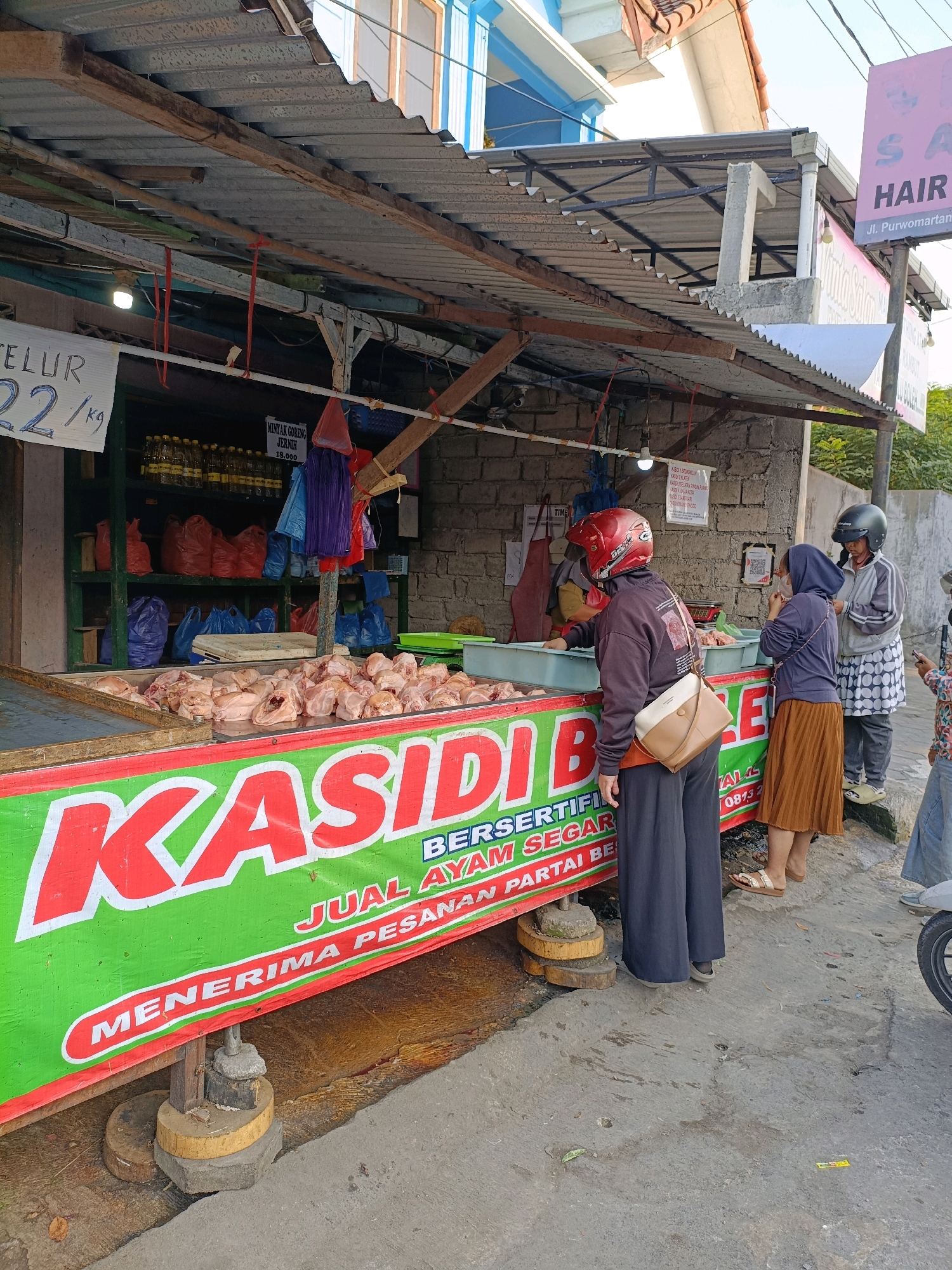 The image depicts an outdoor food stall in Purwomartani, Indonesia, specializing in the sale of raw chicken. A bright green banner with red lettering spans the front of the stall, prominently displaying "KASIDI B..." and further text in smaller font reading "BERSERTIFIKASI JUAL AYAM SEGAR MENERIMA PESANAN PARTAI BESAR" (Certified Sale of Fresh Chicken, Accepts Wholesale Orders). Beneath the banner, a large display case is filled with an abundance of raw, cut chicken pieces. Behind the stall, shelves are stocked with bottles of cooking oil and various bags, some red and some blue, suggesting packaged goods or other merchandise. The stall is covered by a corrugated metal roof supported by wooden beams and metal framing. A brick wall forms the back of the stall.
Three individuals are visible interacting with the stall. One person, wearing a red helmet and a dark purple long-sleeved shirt with a tan and cream crossbody bag, appears to be interacting with the stall owner, whose hand is visible reaching into the chicken display. Another person, wearing a blue headscarf and a brown pleated skirt, stands to the right, looking towards the stall. A third person, wearing a grey jacket and a black helmet, stands behind the first two, observing. The ground in front of the stall is paved with concrete. In the background, a building with blue and white elements is visible, along with a sign for "SA HAIR," indicating a nearby salon. The scene suggests a typical street-side market environment.