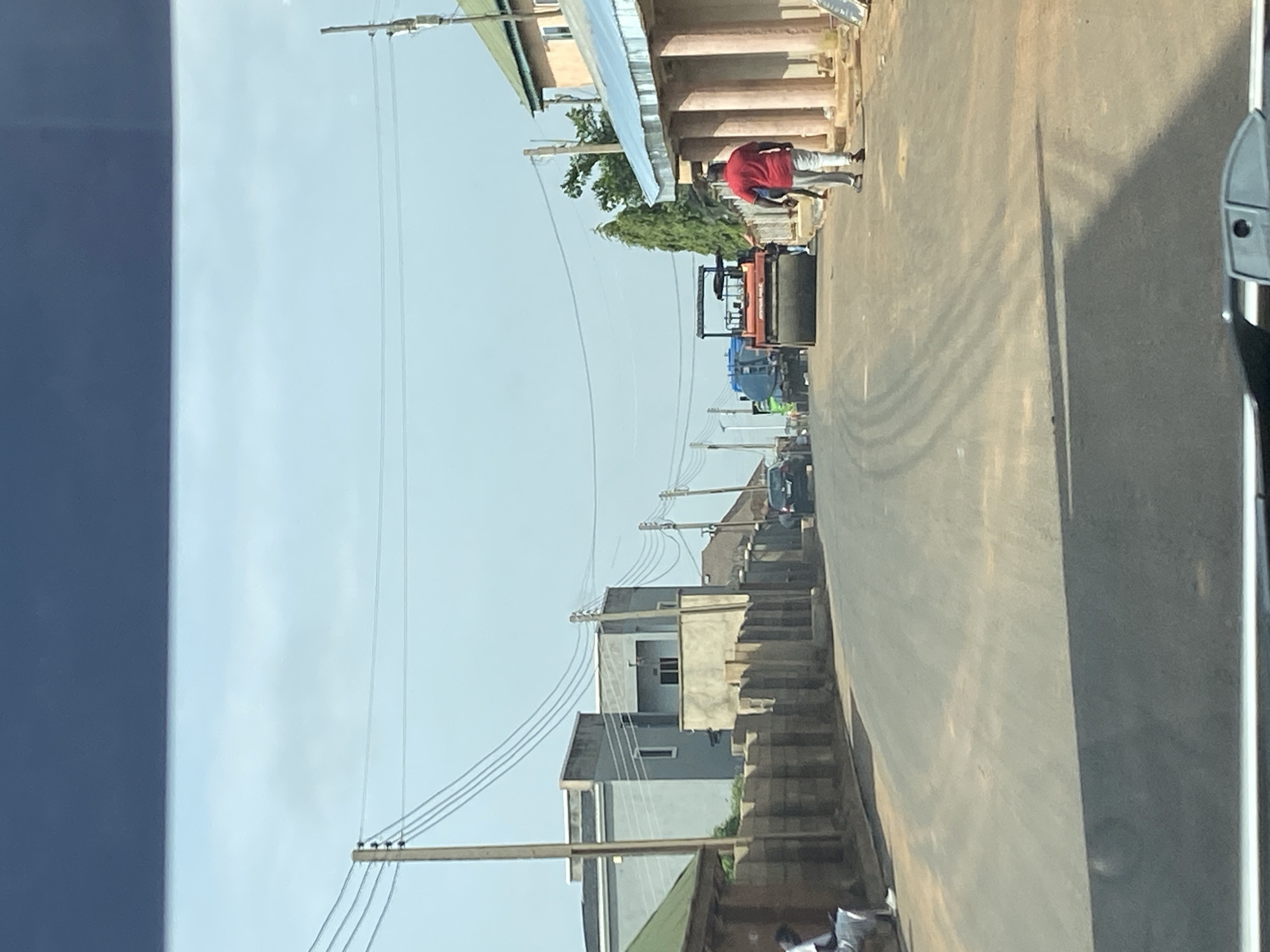 The image shows a street scene in Bar Jirgi Summa, Nigeria. The street is paved and appears to be undergoing construction or maintenance, as indicated by the presence of a large road roller in the middle of the road. Several vehicles, including a black car and a large blue and green truck, are visible on the road. Two men are walking in the street, one wearing a red shirt and white pants, and another in dark clothing. Buildings line both sides of the street, some appearing to be residential homes with concrete block walls and metal roofs, while others are multi-story structures. Overhead power lines are present, supported by wooden utility poles. The sky is clear and blue. The scene suggests an active urban or semi-urban environment.