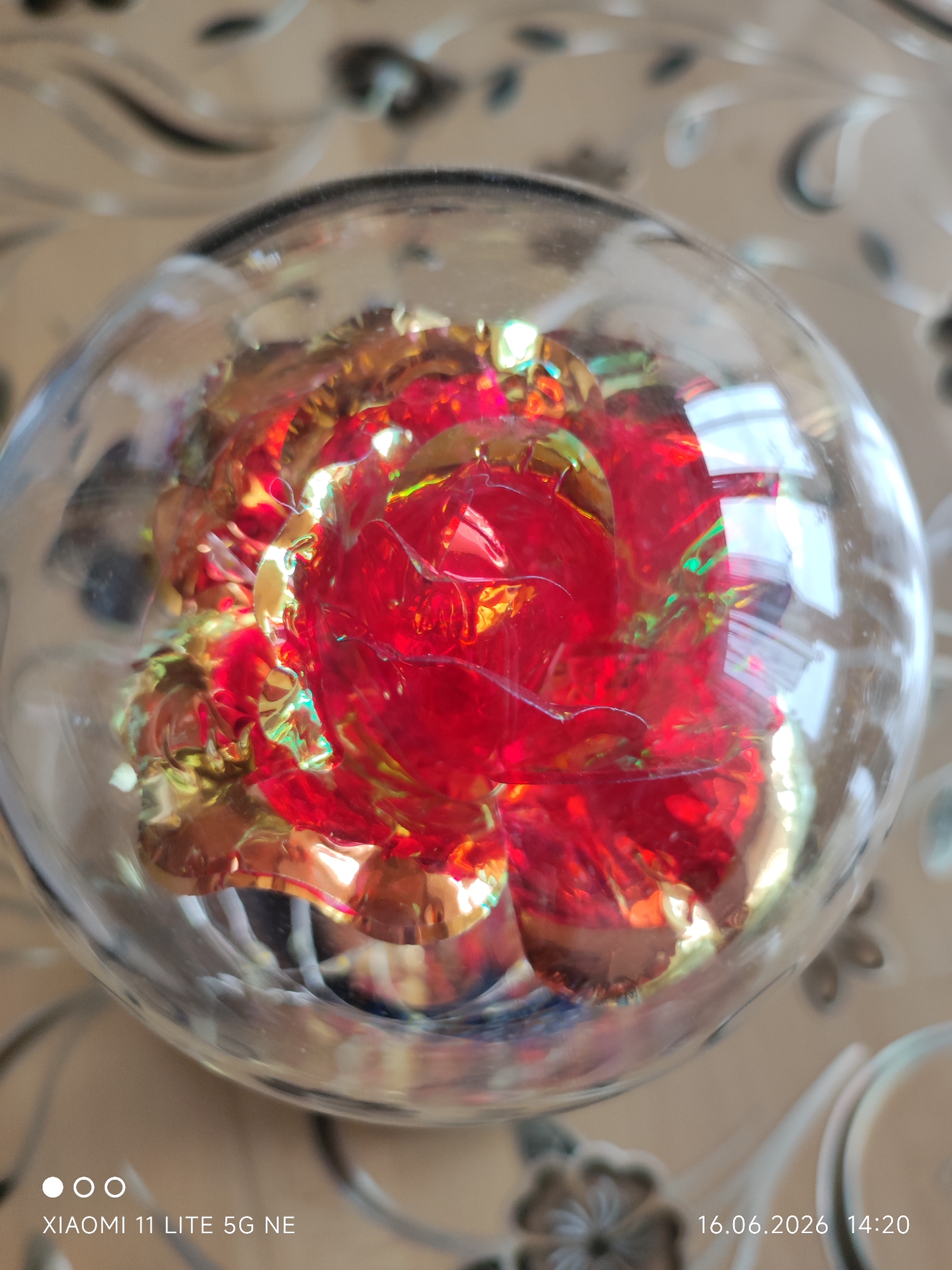 This is a close-up, overhead shot of a red, iridescent rose encased in a clear glass orb, possibly a paperweight or decorative item. The rose appears to be made of a metallic or plastic material, with layers of petals that catch the light, creating vibrant red and golden hues with hints of green. The glass orb has a smooth, spherical surface that slightly distorts the view of the rose within. The background is out of focus but appears to be a textured surface, possibly a table with a patterned tablecloth or placemat in neutral tones of beige and brown. The image also contains text overlays, indicating "XIAOMI 11 LITE 5G NE" in the bottom left corner and "16.06.2026 14:20" in the bottom right corner. There are no people or discernible actions taking place. The scene is static and focused on the ornate rose within the glass orb. There is no information to indicate the location in Böblingen, Germany.