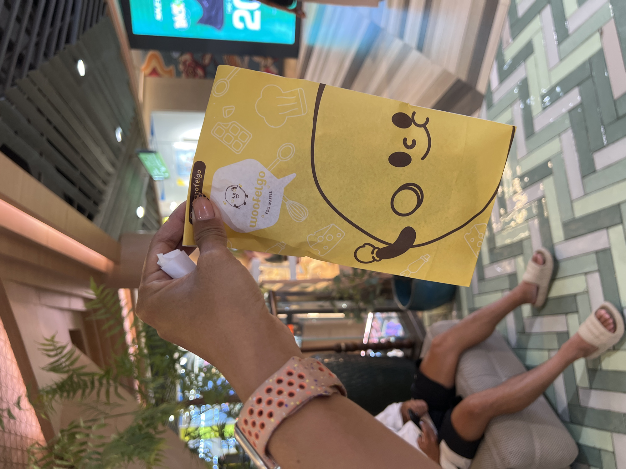 An individual's hand holds a yellow paper packaging with cartoon graphics and text that reads "woofelfgo EGG WAFFLE" in front of a blurred background. The packaging features a character with large eyes and a smiling mouth, along with illustrations of cooking utensils and a chocolate bar. The background shows a person sitting on a light-colored couch, wearing shorts and sandals. The environment appears to be an indoor public space, possibly a mall, with a patterned floor and some greenery. The specific location in Denpasar, Indonesia, is not discernible from the image itself.