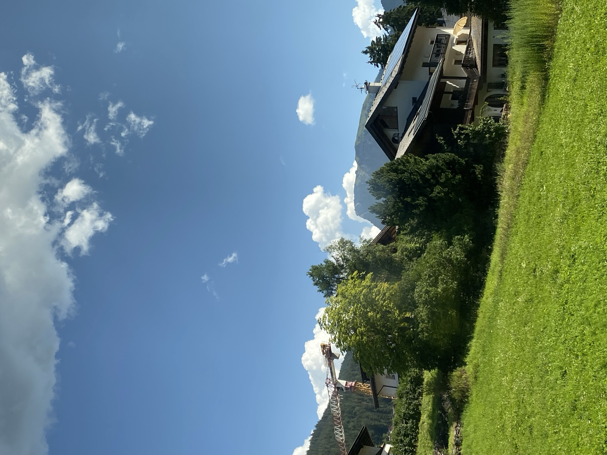 This image depicts a sunny day in the mountainous region of Urtijëi - St. Ulrich in Gröden - Ortisei, Italy. The scene is dominated by a vibrant green meadow in the foreground, gently sloping upwards towards a cluster of buildings partially obscured by lush trees. In the mid-ground, several houses with dark-tiled roofs and wooden balconies are visible. One house stands out with its white facade and dark wood accents, featuring solar panels on its roof. Adjacent to this, a large construction crane with yellow and red markings is positioned, suggesting ongoing development or renovation in the area.
The background is characterized by a dense forest covering a mountainside, with taller, darker green coniferous trees towards the left and a lighter green, more rounded peak in the distance on the right, partially shrouded in clouds. The sky is a clear, bright blue, punctuated by scattered white clouds, some of which are large and puffy, while others are wispy. The overall impression is one of a serene, rural mountain landscape with evidence of human habitation and construction. No people are visible in the image.