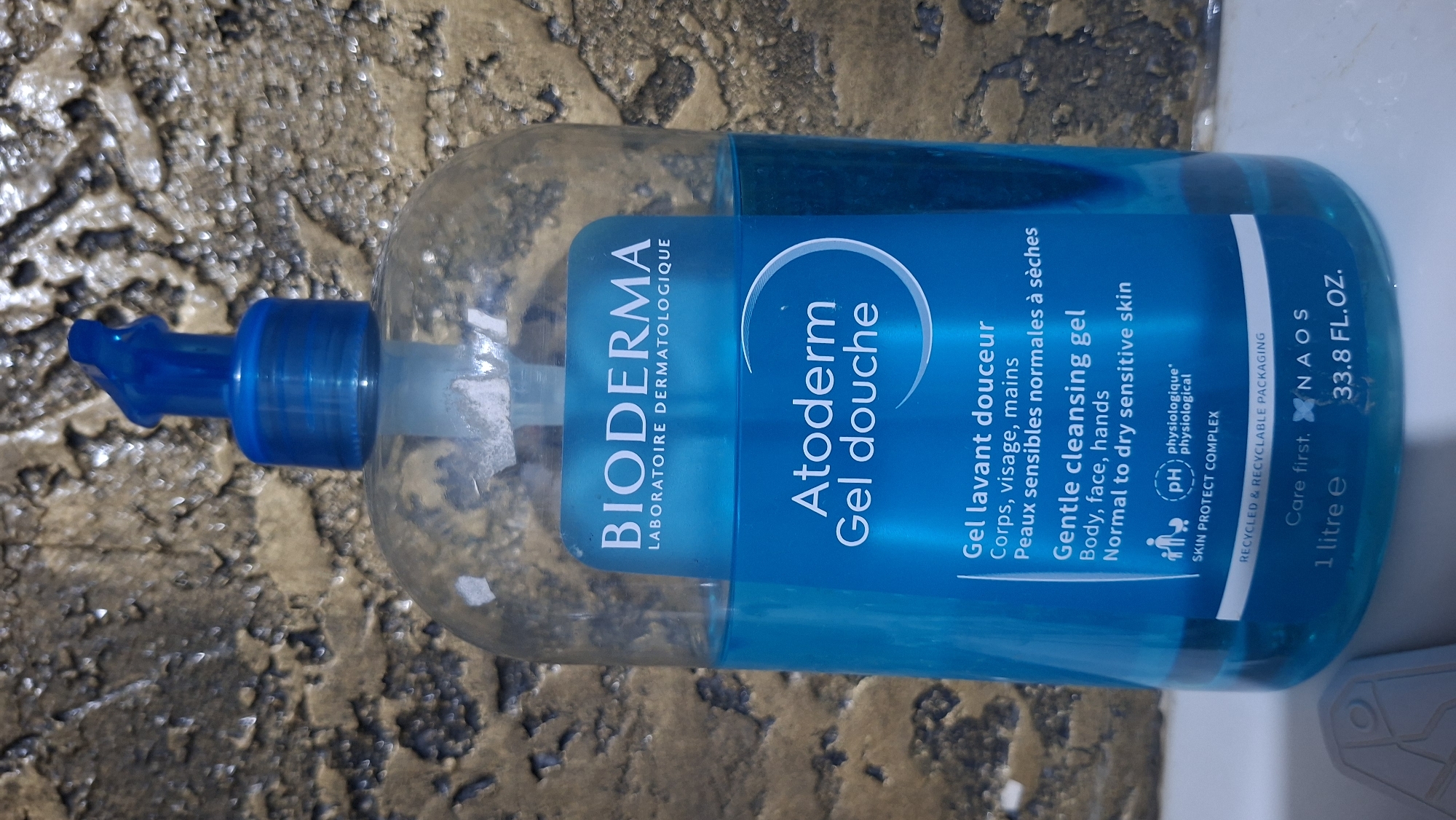 A clear plastic bottle with a blue pump dispenser is filled with blue liquid. The bottle is labeled with "BIODERMA LABORATOIRE DERMATOLOGIQUE" at the top, followed by "Atoderm Gel douche" and "Gentle cleansing gel" in English. Below that are more product details in French and English, including "Corps, visage, mains" and "Body, face, hands" and "Peaux sensibles normales à sèches" and "Normal to dry sensitive skin." There is also a "pH physiologique" symbol and "SKIN PROTECT COMPLEX." The bottom of the label shows "RECYCLED & RECYCLABLE PACKAGING," "Care first. NAOS," "1 litre e," and "33.8 FL.OZ." The bottle is set against a textured, mottled brown and beige background that resembles a wall. The image is cropped to focus on the bottle and its label. There are no people or any indication of a specific location in the city of Voinova, Moldova.