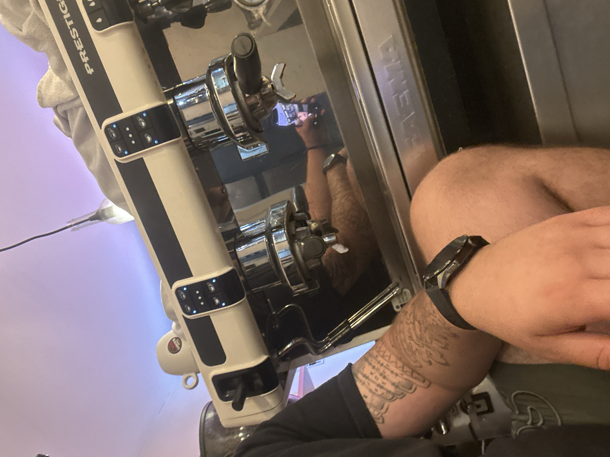 This close-up, tilted image captures a person's lower body and arm next to a professional espresso machine, likely in an indoor setting such as a cafe or kitchen in Lom, Bulgaria.
The main subjects are the person and the coffee machine. The person's left arm is prominently featured, displaying a tattoo that resembles angel wings or feathers along with some script. A dark-colored watch or fitness tracker is worn on the wrist. Part of a bare leg or thigh is also visible in the foreground. The person appears to be seated or leaning near the machine.
The coffee machine is a multi-group espresso maker, primarily cream or off-white with distinct dark vertical stripes. The brand name "PRESTIGE" is clearly visible running vertically along the side. Two group heads with chrome finishes and control panels are present, featuring multiple buttons with active blue indicator lights. A reflection on the machine's shiny metallic surface shows a hand holding a smartphone, suggesting the photo was taken as a selfie or by the subject themselves. Faintly visible on another metallic panel is the word "BREVA".
The background exhibits a soft, diffused purple or lavender light, indicating artificial illumination. The overall scene suggests a casual moment, perhaps during work or a break, at an establishment equipped with a high-quality coffee machine. No specific emotions are discernible.