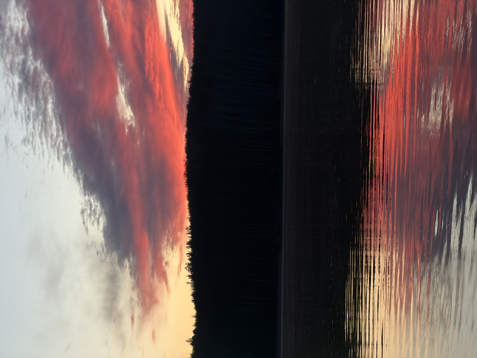 The image depicts a sunset reflected on a body of water, likely a lake or river. The location is implied to be in or near Chaikovsky, Russia. The sky is a mix of colors, including reds, oranges, whites and grays, indicative of sunset or sunrise. The right side of the image shows the reflections of these colors in the water. A dark, silhouetted line of trees or forest separates the sky from the water's reflection. The water is dark, with vertical lines suggesting movement or ripples. There are no people or objects clearly visible in the image.