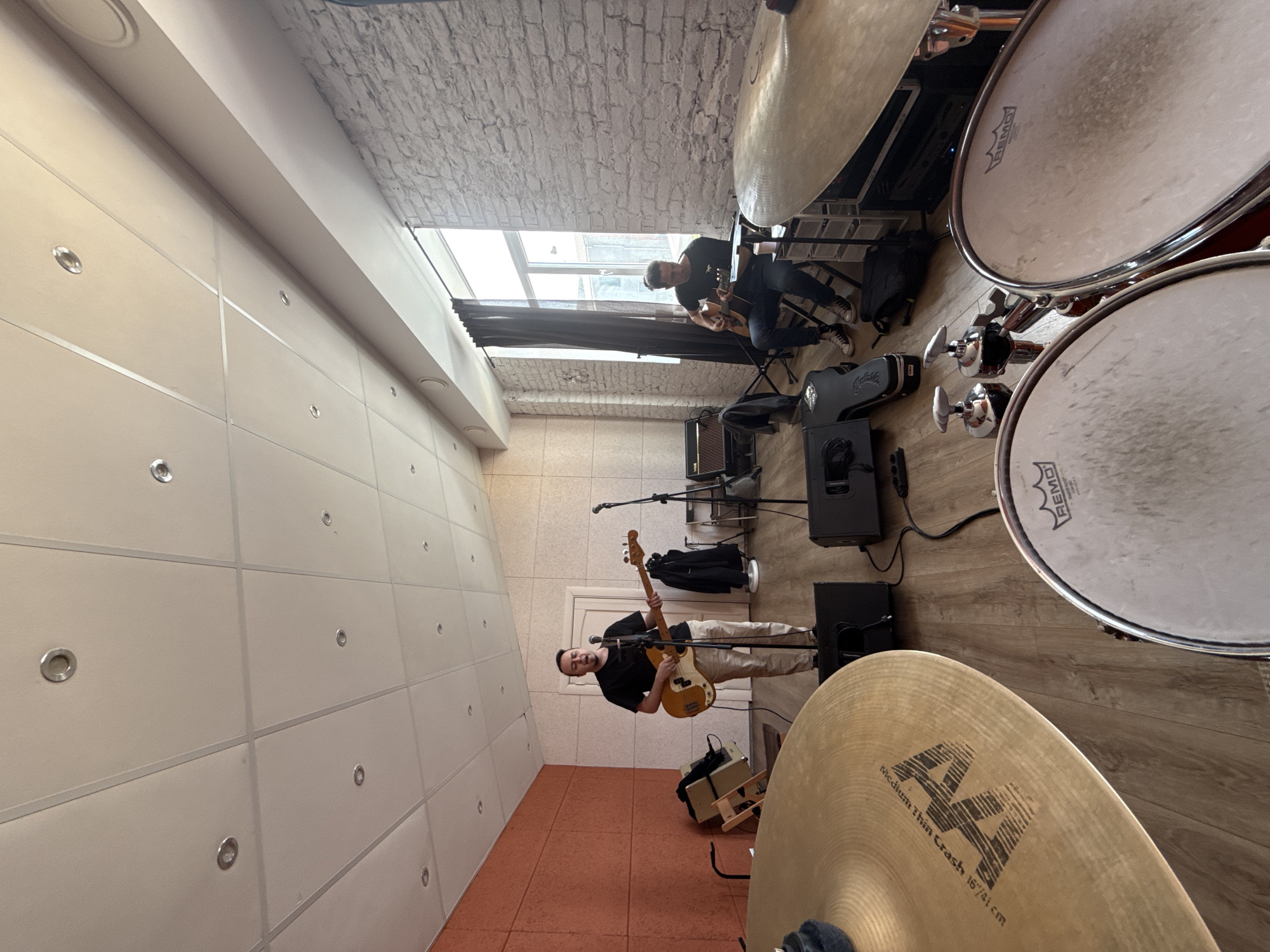 The image shows a music rehearsal room. Two musicians are present. One, wearing a black t-shirt and light-colored pants, is playing a bass guitar. He stands near a microphone on a stand. The second musician is seated and plays an electric guitar. The room contains a drum set with Remo drumheads and a Sabian AA Medium Thin Crash cymbal. Musical equipment, including amplifiers and stands, are scattered throughout the space. The walls are tiled. The location is likely a music studio or practice space, possibly in Moscow, Russia. A window is visible in the background, with dark curtains.