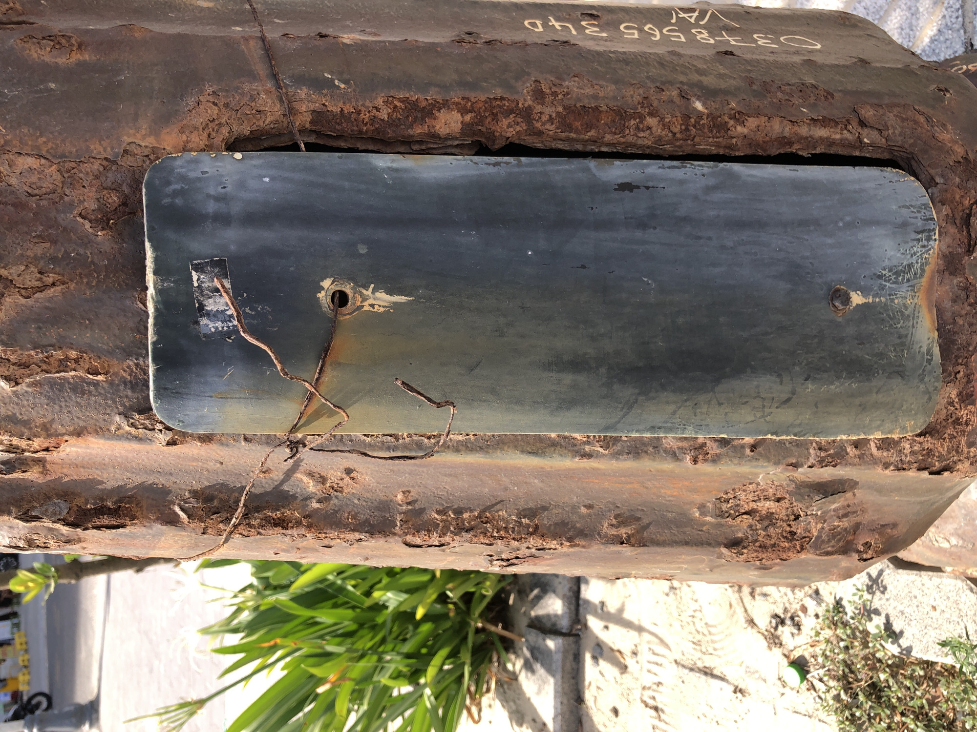The image depicts a close-up of a heavily rusted metal pole in Da Nang, Vietnam. A dark, rectangular plate is attached to the pole with thin, rusty wires and a visible screw. The pole itself is severely corroded, with large flakes of rust and an uneven, pitted surface. To the left of the pole, green foliage and part of a paved walkway are visible. On the right side of the pole, faint yellow lettering can be seen, possibly a serial number or identification marking, which reads "0378565 340 VA". The overall impression is one of advanced decay and neglect of the metal structure.