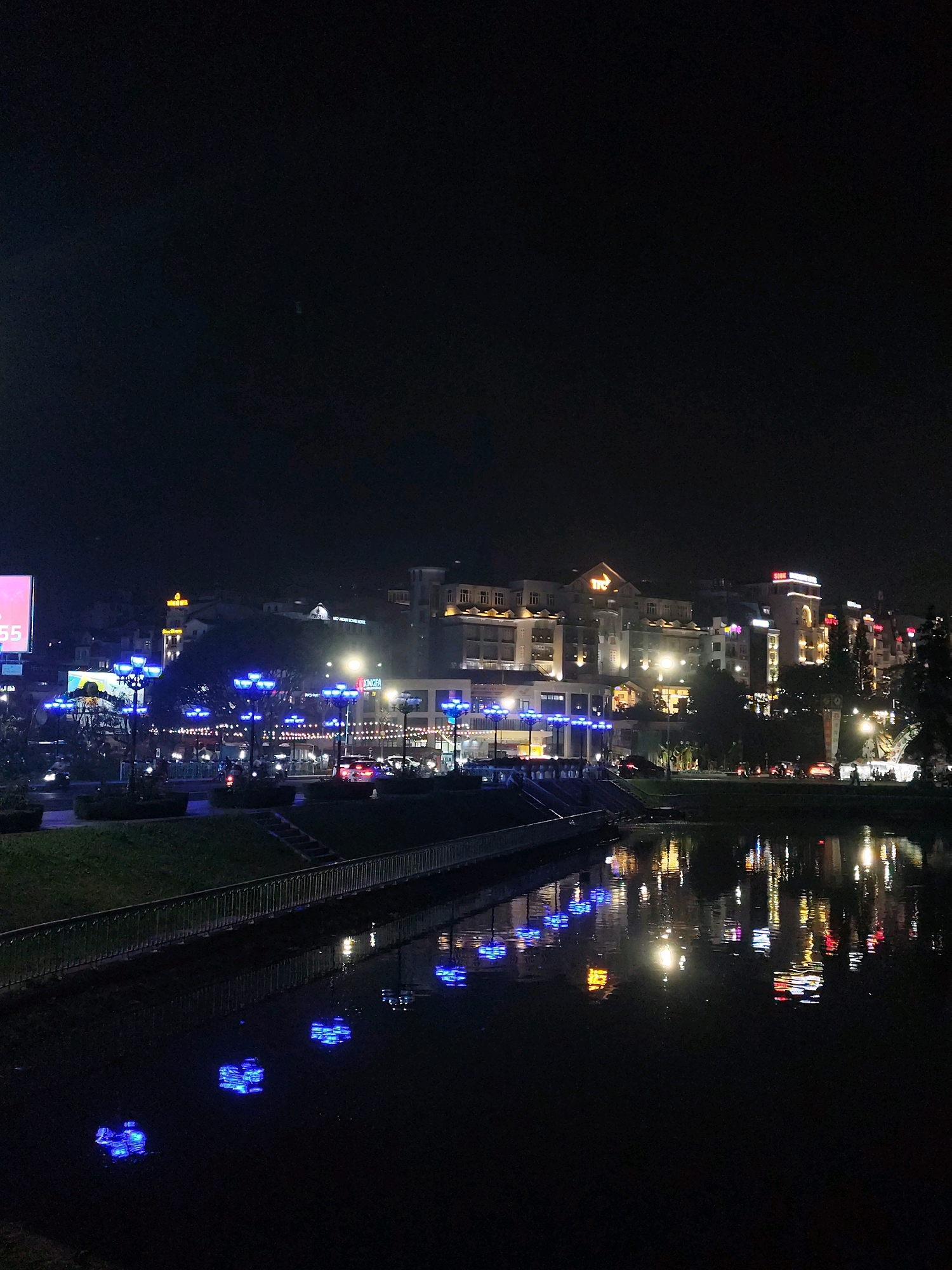 This image captures a nighttime view of Xuan Huong Lake in Da Lat, Vietnam. The city skyline is illuminated with numerous lights from hotels, buildings, and streetlights. Prominent buildings with signs such as "ITC" and "XINGFA HOTEL" are visible. Decorative blue streetlights line the edge of the lake, and their reflections, along with the reflections of other city lights, create vibrant patterns on the dark water. A grassy embankment with a metal railing separates the walkway from the lake. The scene is devoid of any significant human activity, with only faint indications of people in the distance. The overall atmosphere is one of a tranquil, illuminated urban landscape at night.