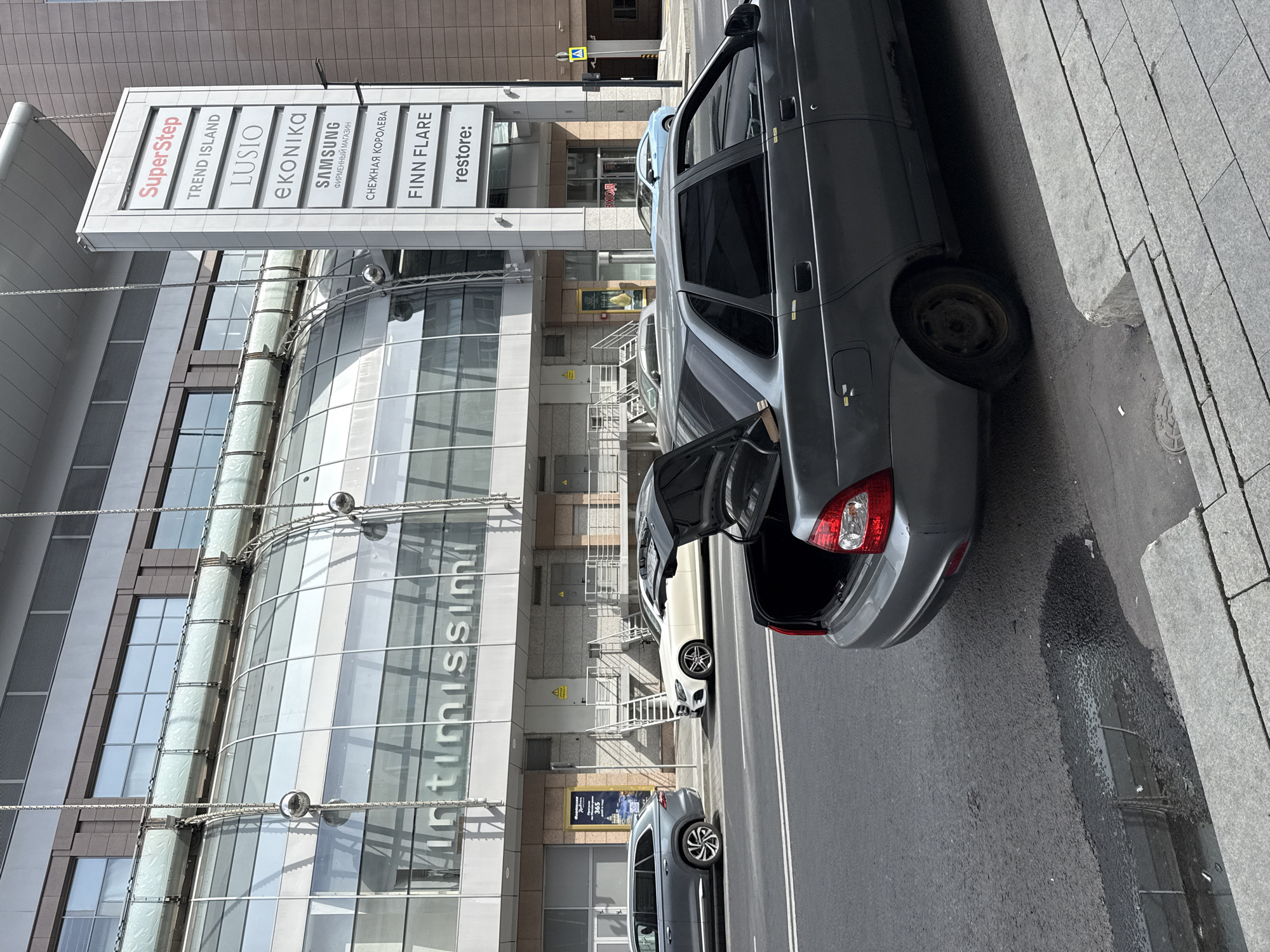 In Moscow, Russia, on a bright, clear day, a grey Lada sedan with its trunk open is parked on the right side of a paved road. The car is facing left, with its rear end closest to the viewer. To the left of the Lada, a white Mercedes-Benz sedan is parked. Further left, a dark grey SUV is parked. In the background, a multi-story building features a large, curved glass facade with the word "intimissimi" visible. Above the glass facade, a vertical white sign displays various brand names, including "SuperStep," "TREND ISLAND," "LUSIO," "eKONIKA," "SAMSUNG," "СНЕЖНАЯ КОРОЛЕВА," "FINN FLARE," and "restore:". A pedestrian crossing signal with a yellow pedestrian figure is visible on the right side of the road. The scene depicts a typical urban street environment with parked vehicles and commercial establishments.