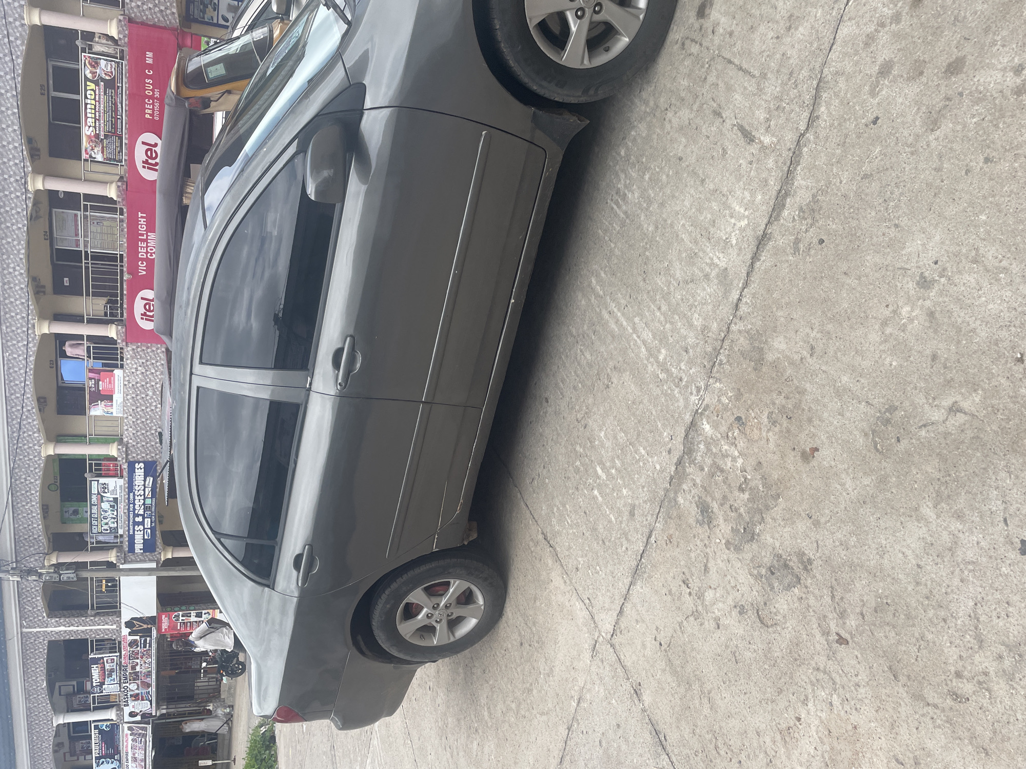This media file depicts a street scene in Jalingo, Nigeria, with a focus on a grey sedan parked on a concrete surface. The car is angled, showing its side profile with the passenger doors visible. The vehicle appears to have some minor damage or rust near the rear wheel arch.
Behind the car, a multi-story building with shops on the ground floor is visible. The building has a light-colored facade with visible signage. Text on the signs includes "Samjoy," "PREC OUS C MM 0701567 301," "itel," "VIC DEE LIGHT COMM," "RICH GIFT GLOBAL COMM," "PHONES & ACCESSORIES," "BASKOO EMPIRE," and "ONES HUB." These signs indicate a commercial area with businesses selling various goods, possibly including electronics and accessories.
A yellow three-wheeled vehicle, commonly known as a Keke Napep, is parked in front of the building, partially obscured by the sedan. Further down the street, a person on a motorcycle can be seen. The lighting suggests it is daytime, and the concrete ground indicates an urban or semi-urban environment. The scene appears to be a typical day in a bustling commercial district.