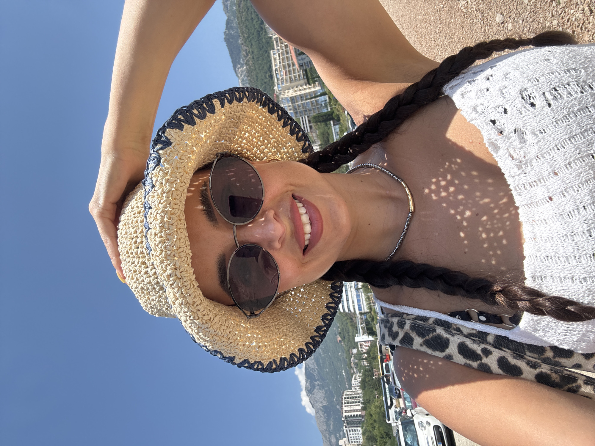 A woman is taking a selfie on a sunny day. She is wearing a straw hat with a blue trim, sunglasses, a white top, and a silver necklace. Her hair is in two braids. In the background, there are buildings, mountains, and cars. The location is Unknown, Unknown.