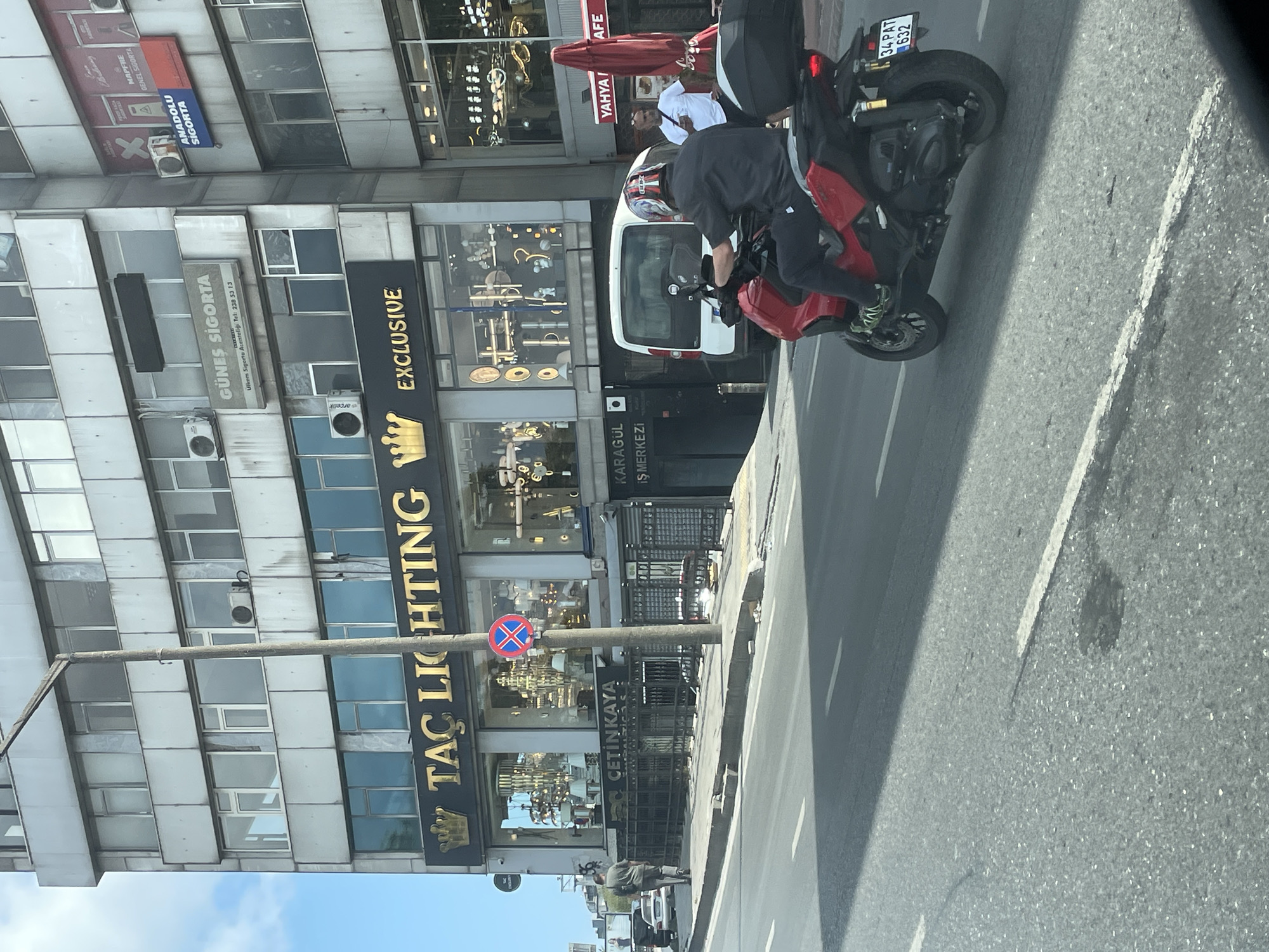 A street-level view captures a red motorcycle in the foreground, ridden by a person wearing a black helmet and black attire, in the city of Beyoğlu, Turkey. The motorcycle is moving on the right side of the road, with its license plate reading "34 PAT 632". Behind the motorcycle, a white van is visible. On the sidewalk, a man in a grey shirt and dark pants walks away from the camera.
The background is dominated by a multi-story building with a light-colored facade. The ground floor features shops with large glass windows displaying lighting fixtures and decor. Prominent signage on the building includes "TAC LIGHTING" and "EXCLUSIVE" in gold lettering. Above these, signs for "GÜNEŞ SİGORTA" and "ANADOLU SİGORTA" are visible. The street is paved with asphalt, and a pedestrian walkway with a metal fence runs alongside the building. Red umbrellas and tables suggest an outdoor seating area for a cafe. The sky is partly cloudy.
