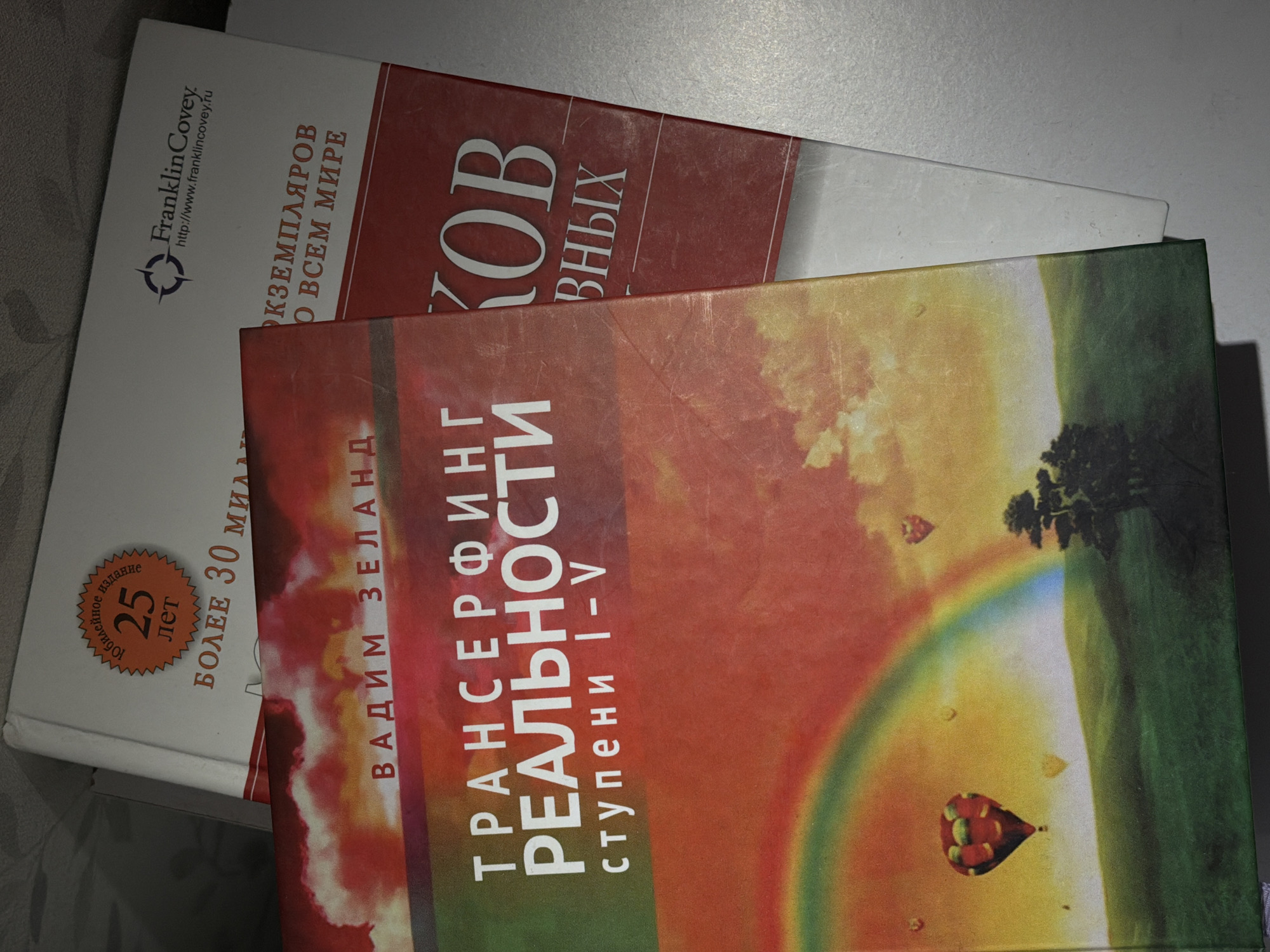 Three books are displayed on a flat surface, likely a table, with two clearly visible and one partially obscured. The image is taken at a diagonal angle, showing the covers of the top two books.
The top book has a white cover. Its upper left corner features the "FranklinCovey." logo and the website "http://www.franklincovey.ru". Text on the left side, partially visible, reads "Юбилейное издание 25 лет" (Anniversary edition 25 years) and "БОЛЕЕ 30 МИЛ" (More than 30 MIL, likely 'million'). Further text on the right side of the cover says "ЭКЗЕМПЛЯРОВ О ВСЕМ МИРЕ" (copies all over the world) and parts of a title: "КОВ ВНЫХ", strongly suggesting "7 НАВЫКОВ ВЫСОКОЭФФЕКТИВНЫХ ЛЮДЕЙ" (The 7 Habits of Highly Effective People).
The middle book has a vibrant, gradient cover transitioning from red and orange to yellow and green. The author's name, "ВАДИМ ЗЕЛАНД" (Vadim Zeland), is prominently displayed vertically on the left. The title, "ТРАНСЕРФИНГ РЕАЛЬНОСТИ" (Reality Transurfing), is written in large white letters, with "ступени I-V" (steps I-V) below it. The cover art depicts a serene landscape with a prominent rainbow arcing across a colorful sky, a dark green cliff or hill on the right, and a small red and yellow hot air balloon floating near the center.
Only a dark spine or edge of a third book is visible beneath the middle book. The lighting suggests an indoor setting. The books show some signs of wear on their covers.