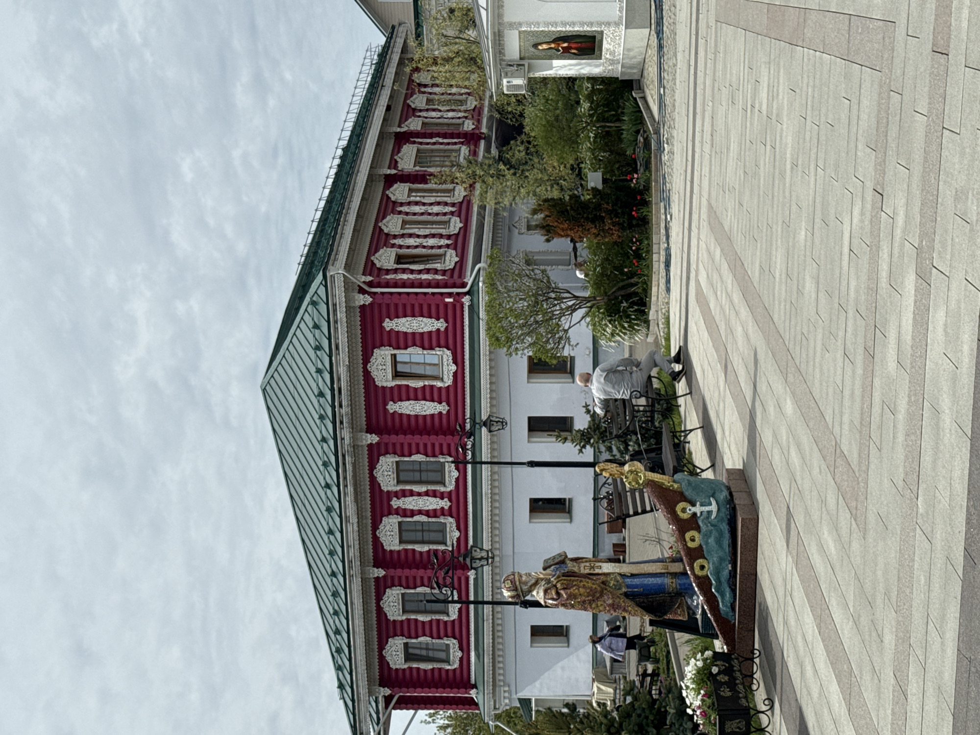 The image shows a view of the Museum of Russian Patriarchate in the city of Arzamas, Russia. A traditional Russian wooden building with red walls and white decorative trim is visible, with a green metal roof. The sky is overcast. The adjacent sidewalk is paved with light gray rectangular stones. Several sculptures and flower arrangements decorate the walkway. A person sits on a bench along the walkway.