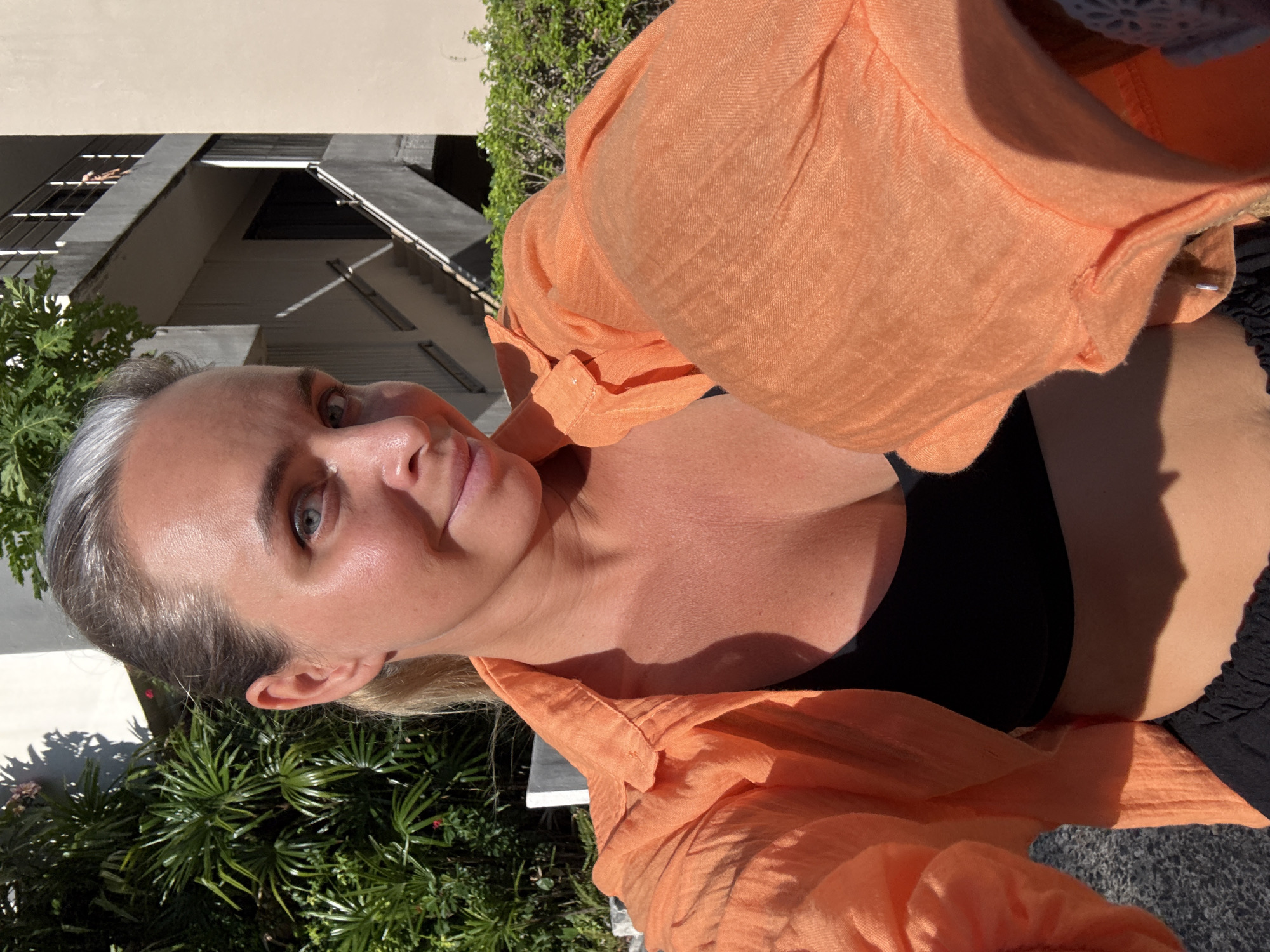 In the city of Ko Samui District, Thailand, a woman is taking a selfie outdoors. She is wearing an unbuttoned orange shirt over a black top and dark shorts. Her hair is pulled back, revealing grey streaks at her temples. The background features lush green foliage and a building with outdoor staircases. The lighting suggests it is a sunny day.